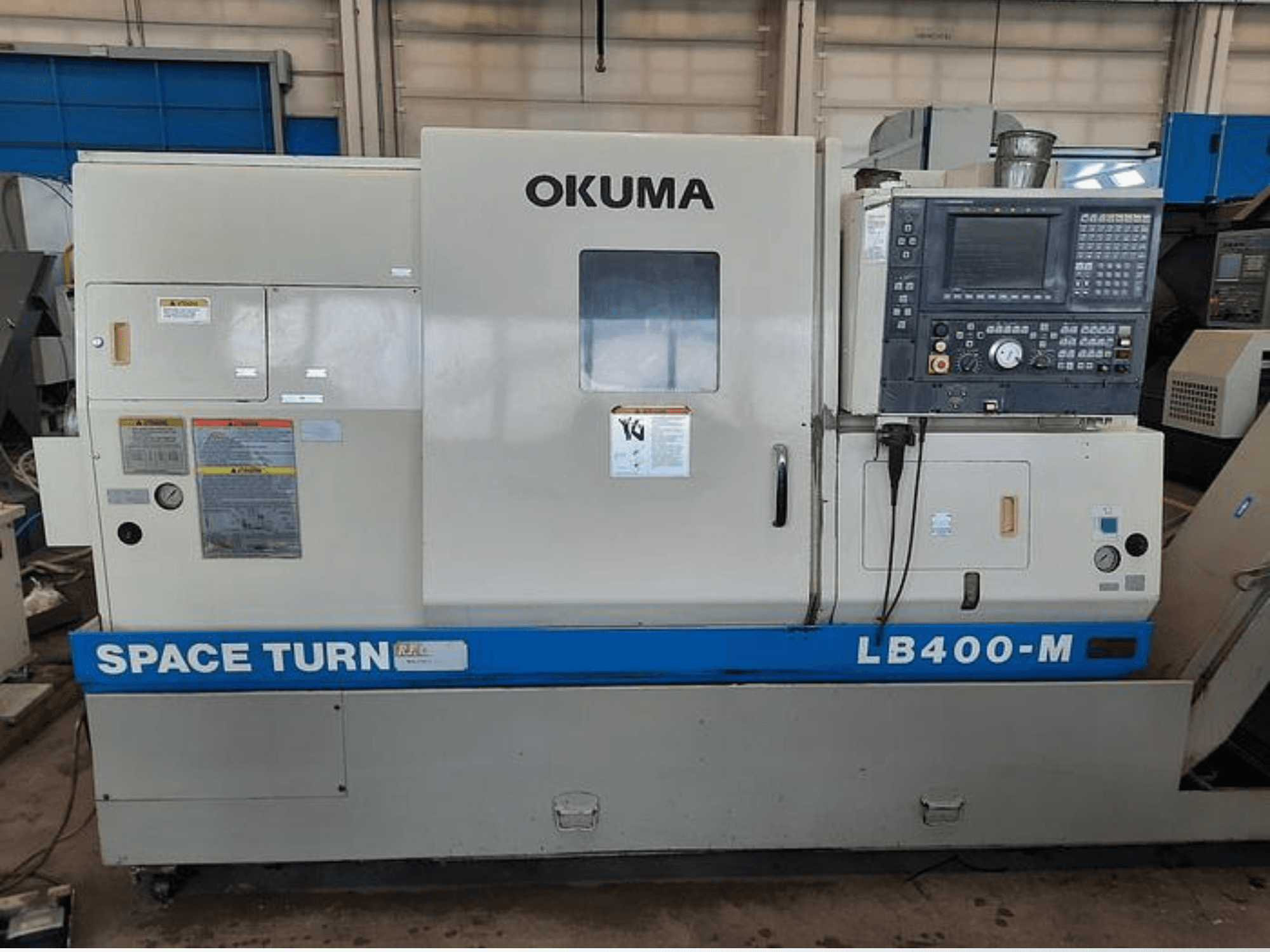 Tokarka CNC Okuma LB400-M, widok z przodu z widocznym panelem sterowania, etykietami ostrzegawczymi i niebieskim oznaczeniem "SPACE TURN".