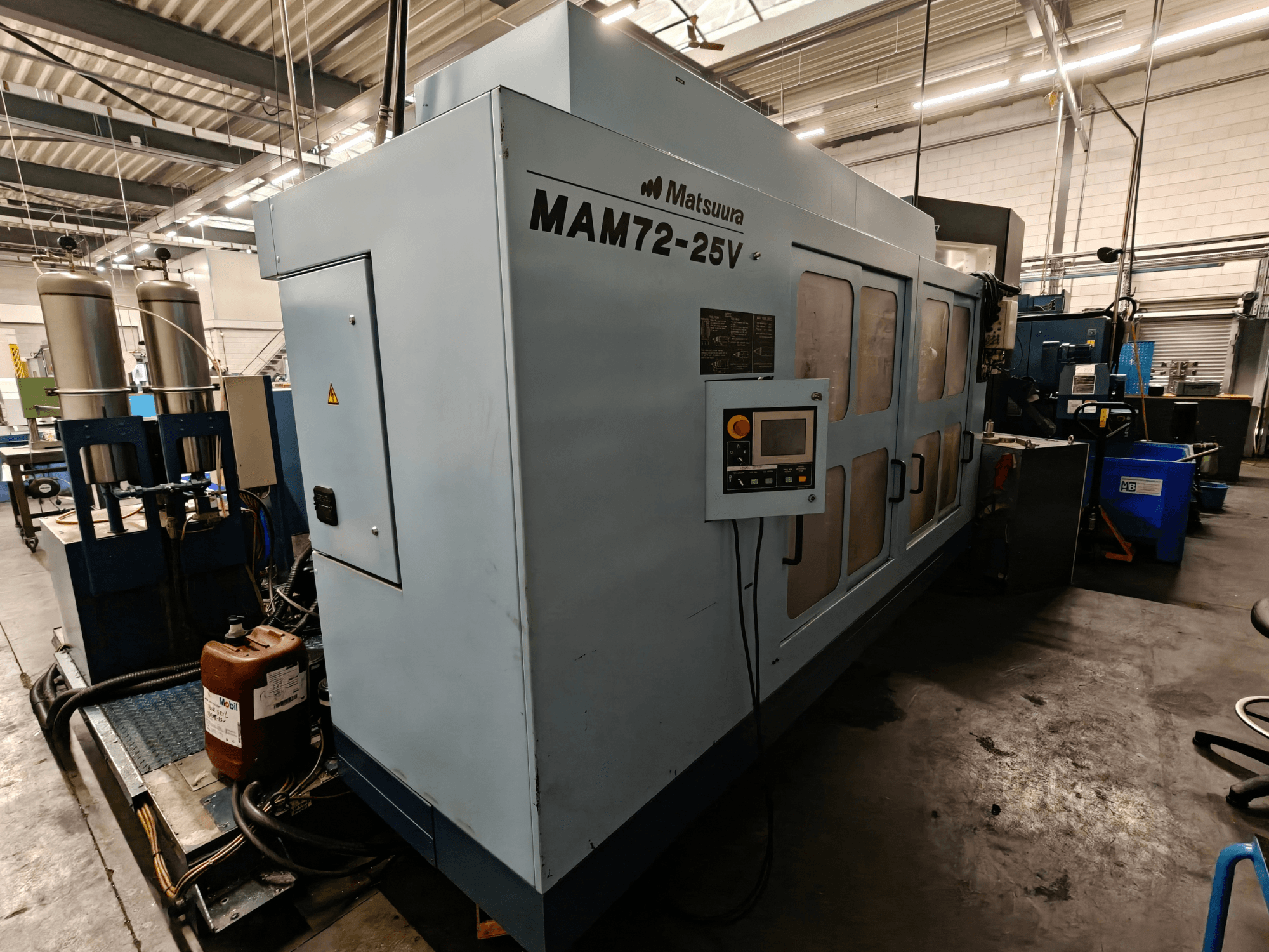 Maszyna CNC Matsuura MAM72-25V, widok z boku, z panelem sterowania, dwoma zbiornikami chłodziwa i przestrzenią roboczą w warunkach przemysłowych.