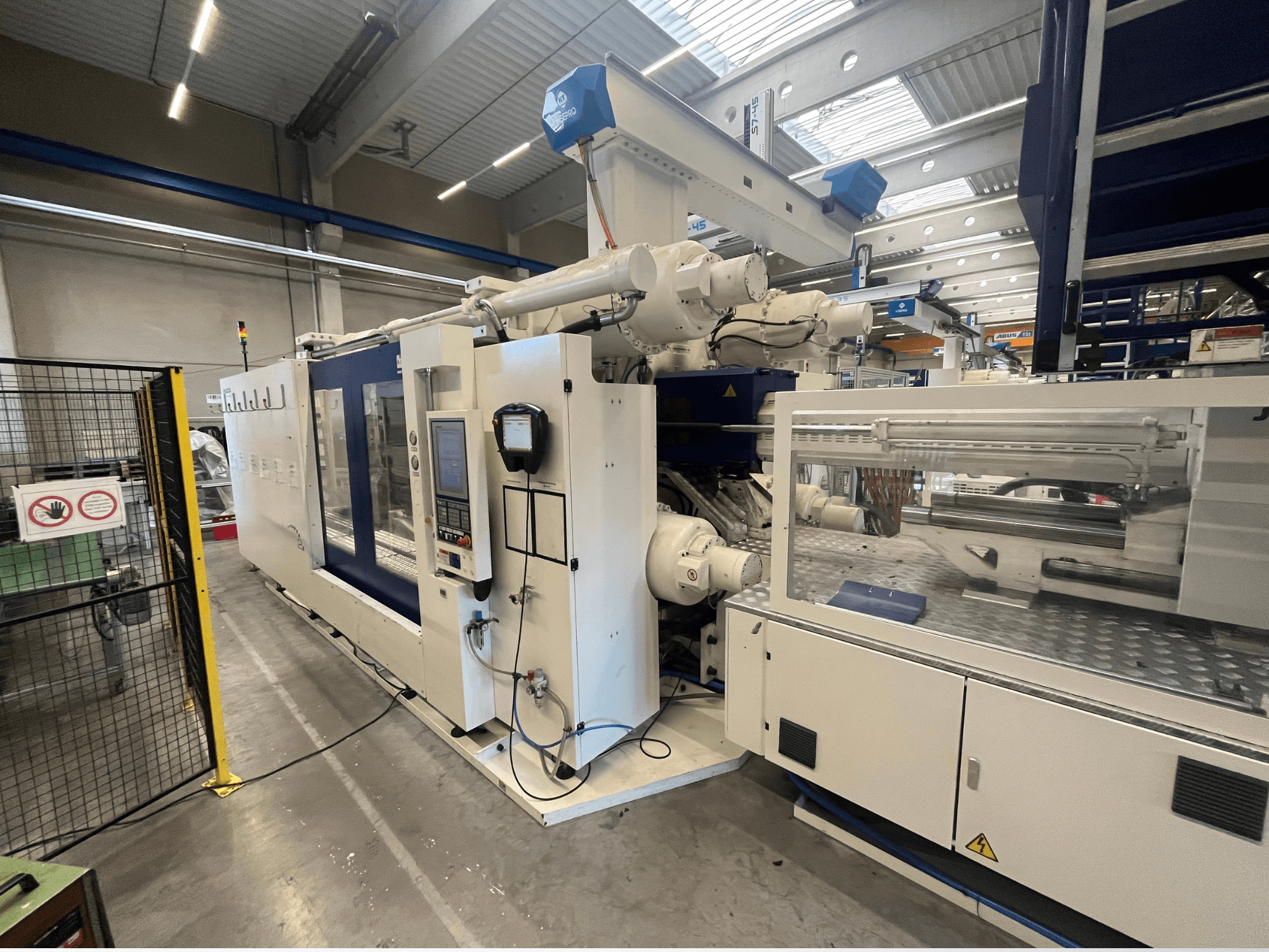 Wtryskarka w kolorze białym i niebieskim, widok z przodu, z panelem sterowania, ramieniem robota i oznakowaniem bezpieczeństwa.