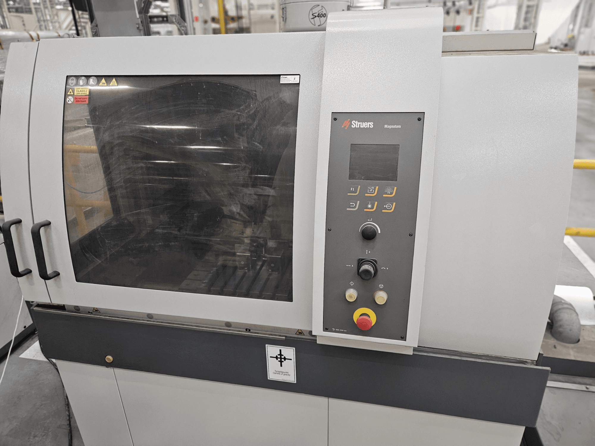Widok z przodu maszyny Struers Magpentor z panelem sterowania, przyciskami i dużym oknem podglądu. Widoczne etykiety techniczne.