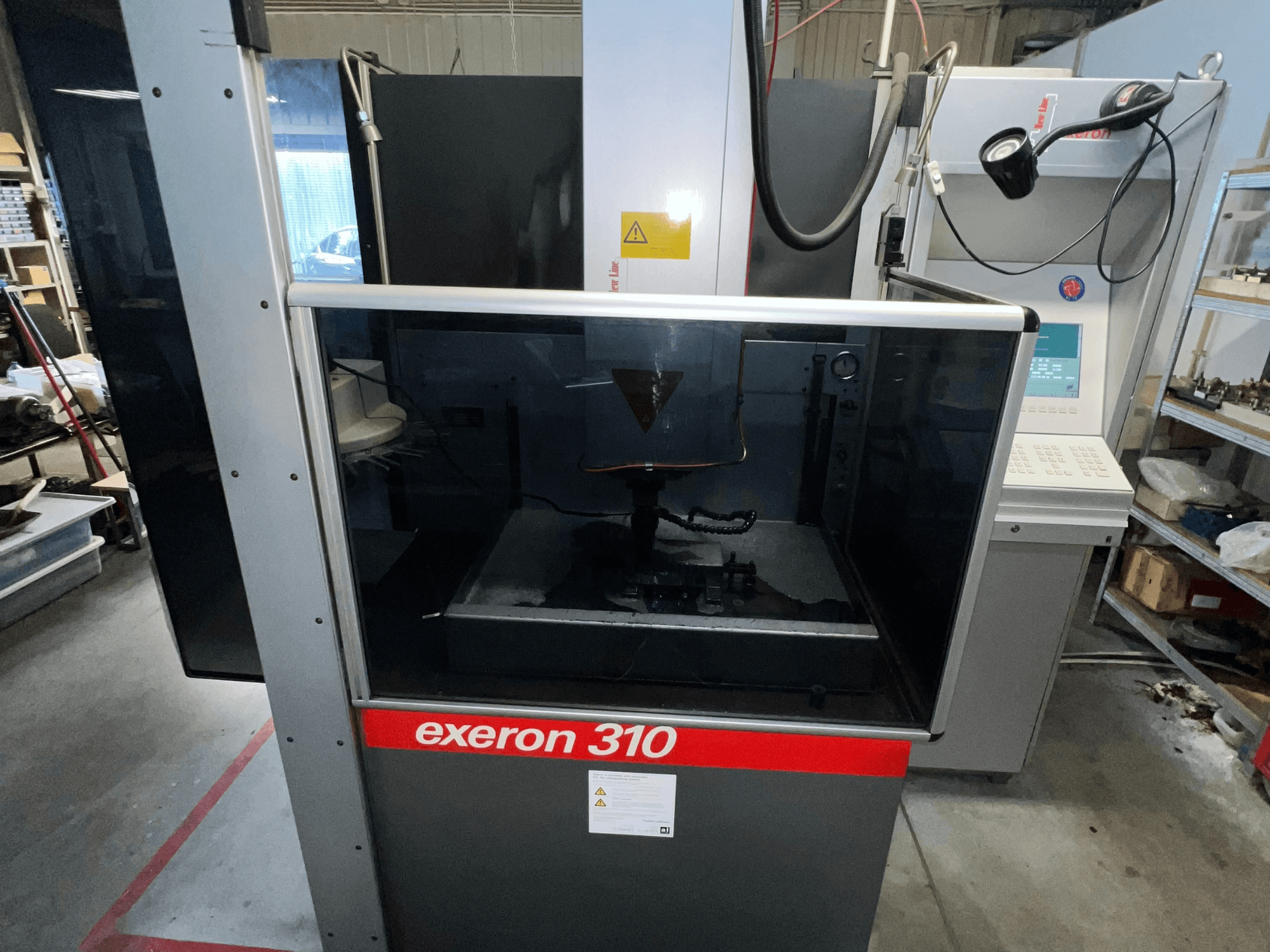 Widok z przodu maszyny Exeron 310, przedstawiający szklaną komorę i panel sterowania wraz z półkami stacji roboczej.