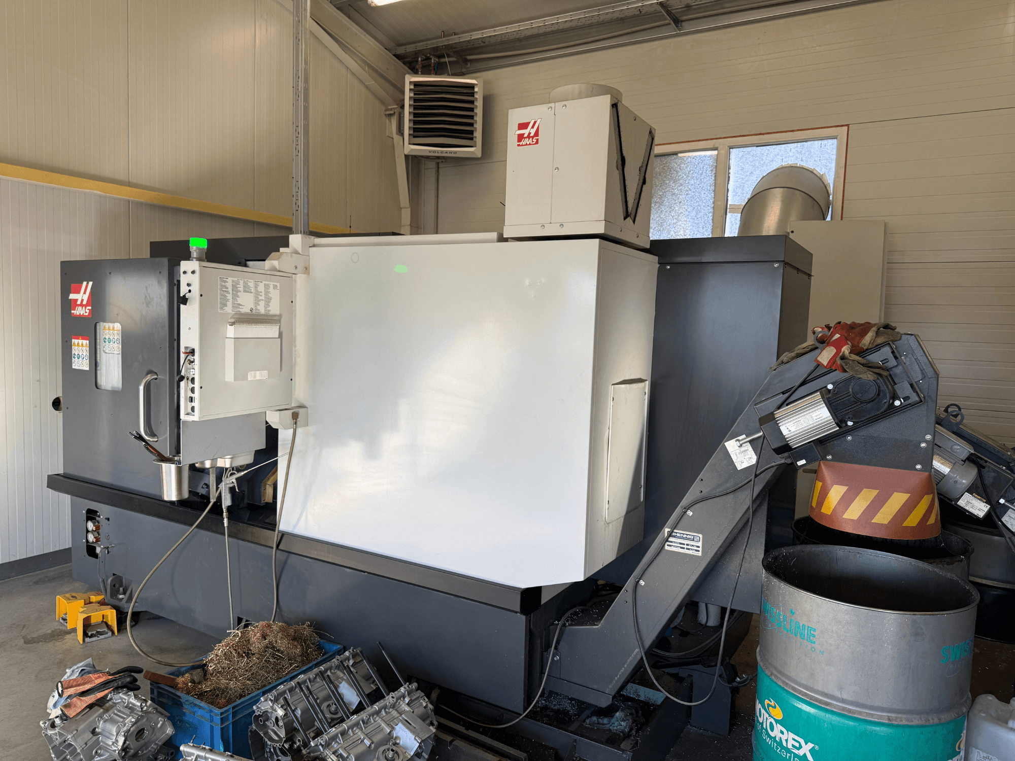 Maszyna CNC Haas z panelem sterowania, widok z boku; widoczne przewody chłodziwa i oleju; złom metalowy w niebieskim pojemniku; otoczenie przemysłowe.