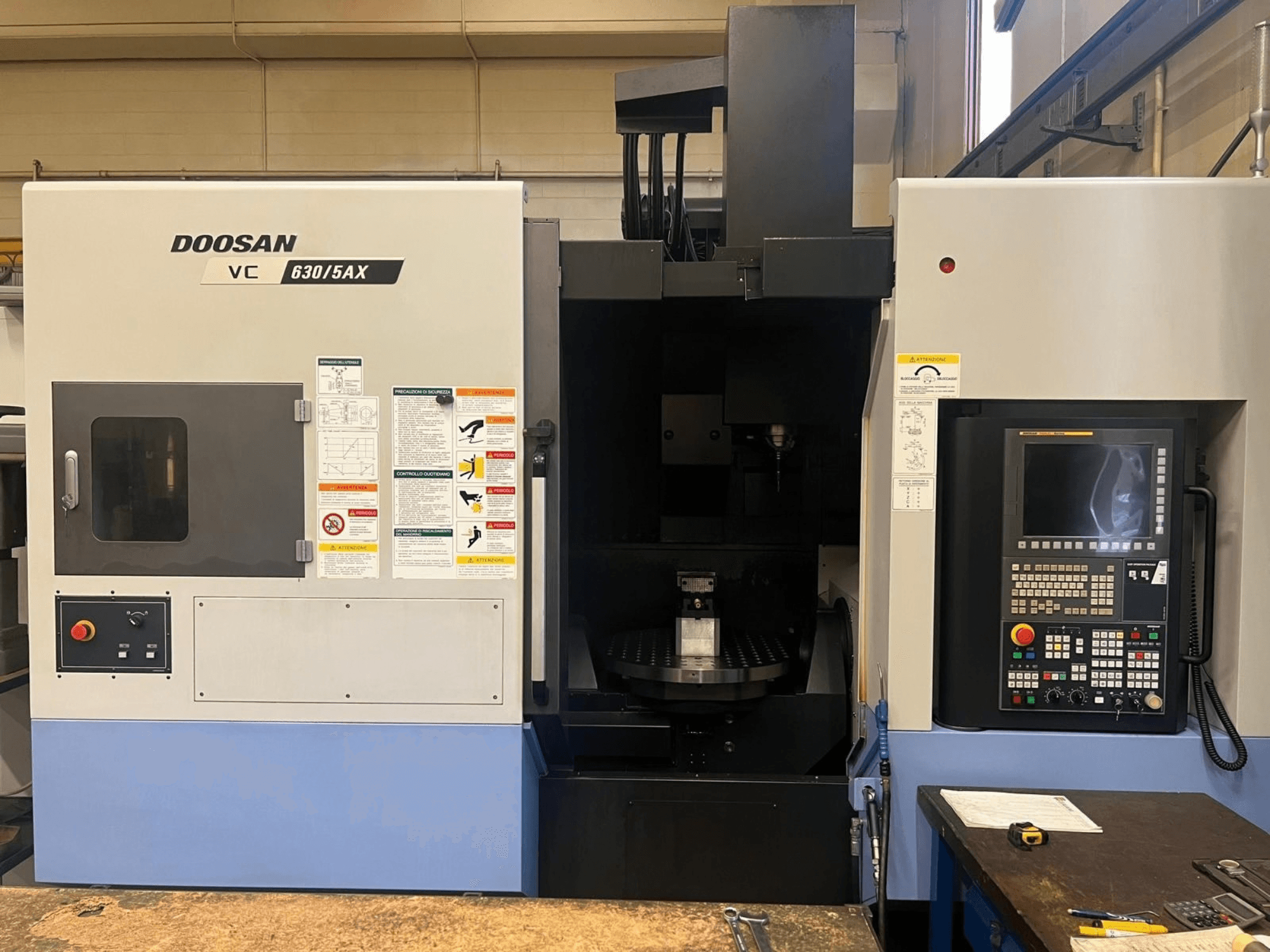 Widok z przodu pionowego centrum obróbczego DOOSAN VC 630/5AX, na którym widoczny jest panel sterowania oraz obszar wrzeciona wraz z etykietami bezpieczeństwa.