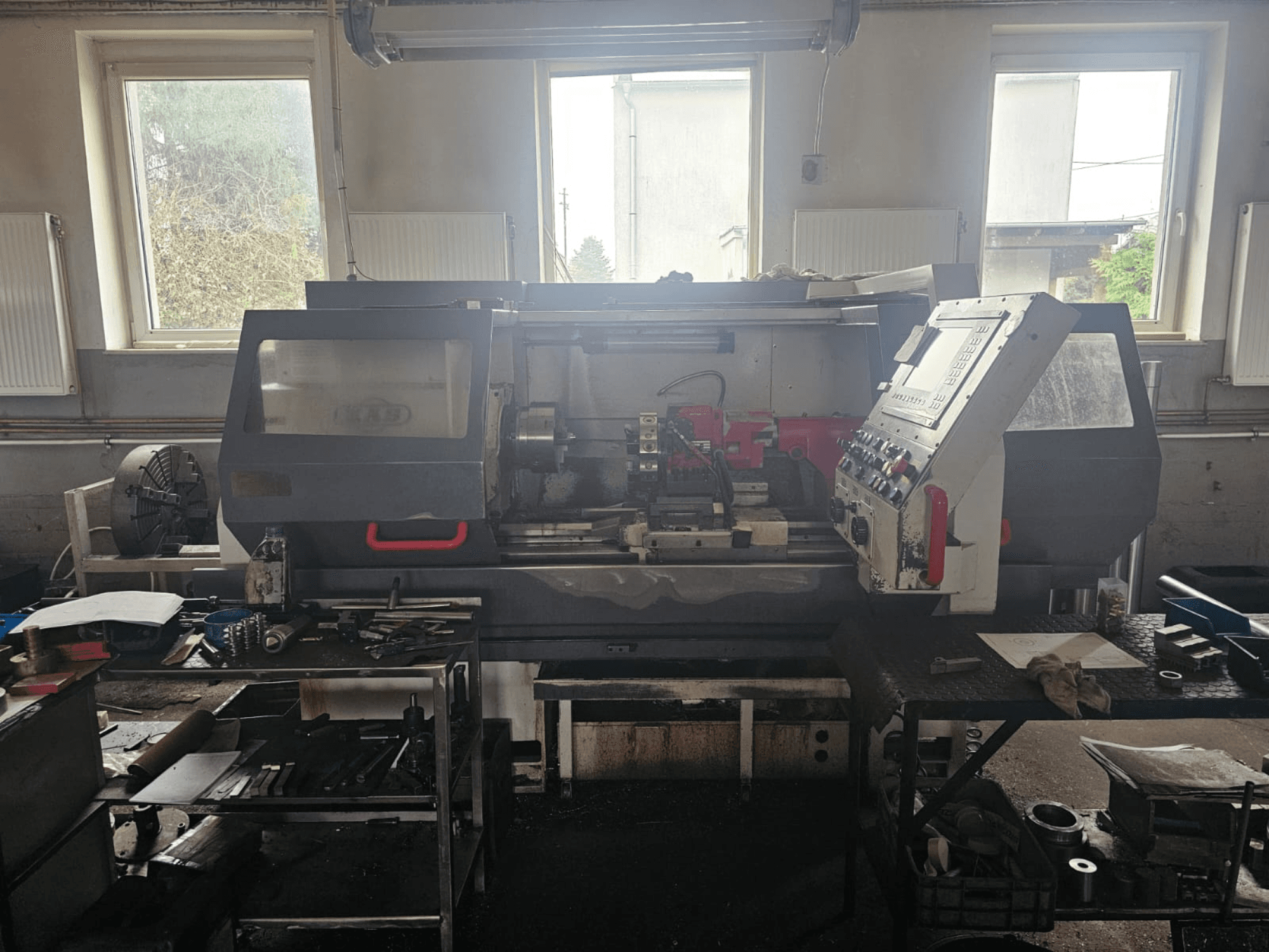 Tokarka w warsztacie, widok z przodu, z panelem sterowania i narzędziami na stołach, otoczona oknami i zagraconą przestrzenią roboczą.