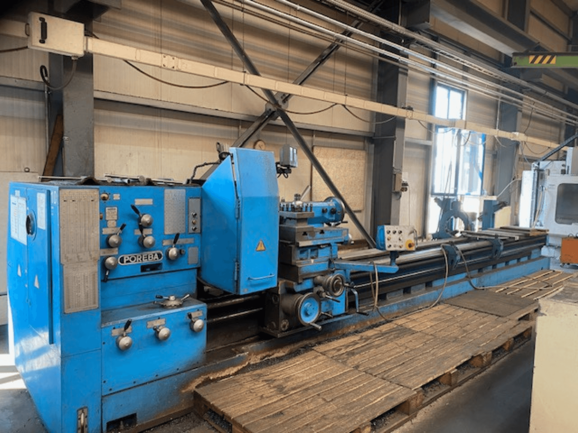 Widok z przodu niebieskiej tokarki POREBA z pokrętłami sterowania, podpórką na narzędzia i drewnianymi paletami pod spodem.