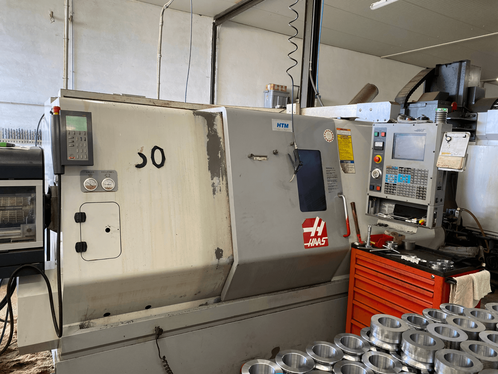 Maszyna CNC Haas z panelem sterowania, widok z przodu, pokazujący przyciski obsługi, mierniki i metalowe części ułożone w pobliżu.