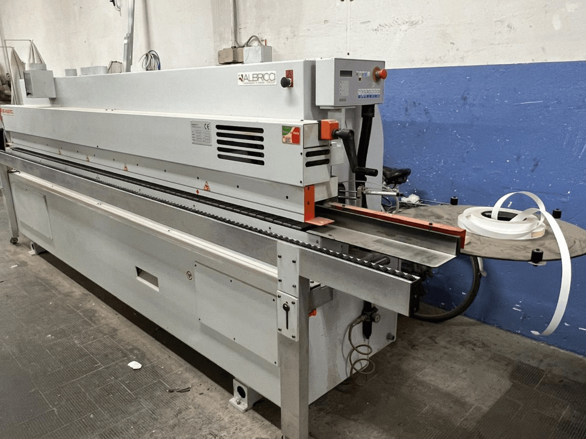 Okleiniarka ALBRICO z widokiem z przodu, z długim przenośnikiem taśmowym i dołączoną rolką taśmy samoprzylepnej.