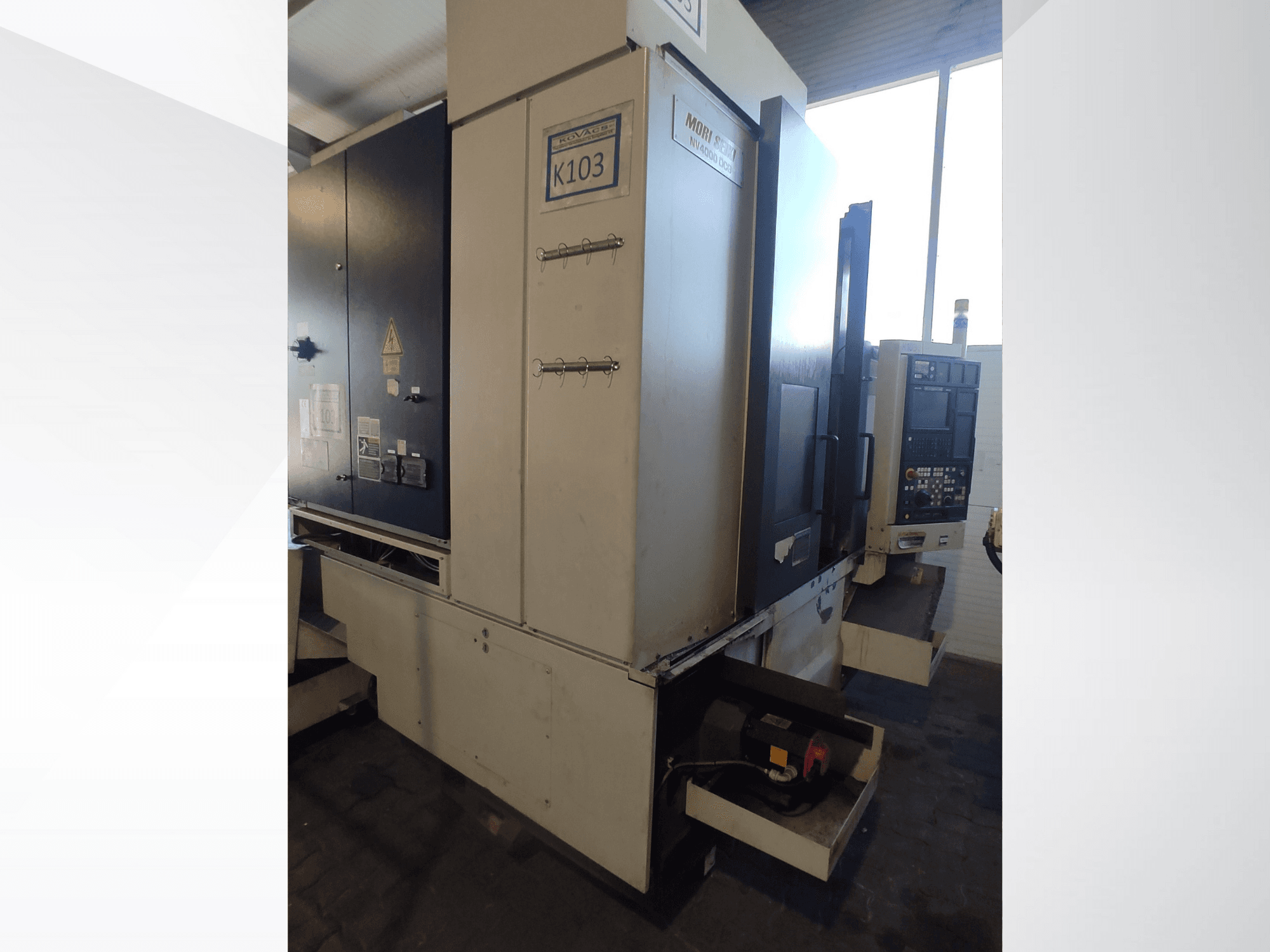 Maszyna CNC Mori Seiki K103 w widoku z przodu, z widocznym panelem sterowania, etykietami bezpieczeństwa i komponentami elektrycznymi.
