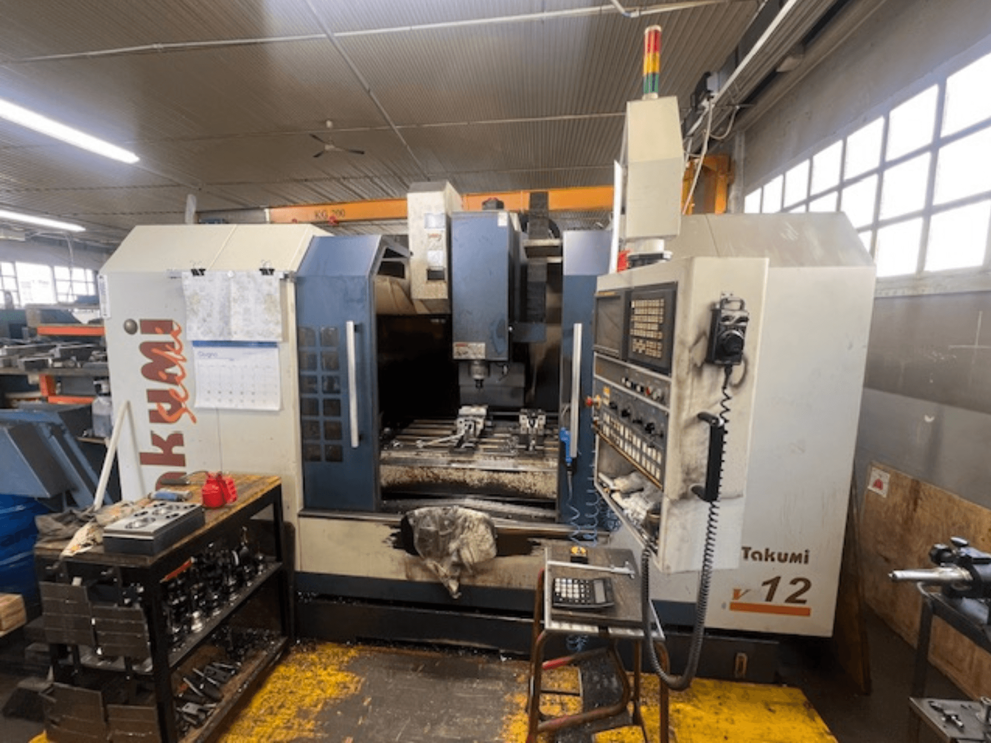 Frezarka CNC Takumi V12 w widoku z boku, pokazująca panel sterowania, obszar cięcia i narzędzia na stacji roboczej.