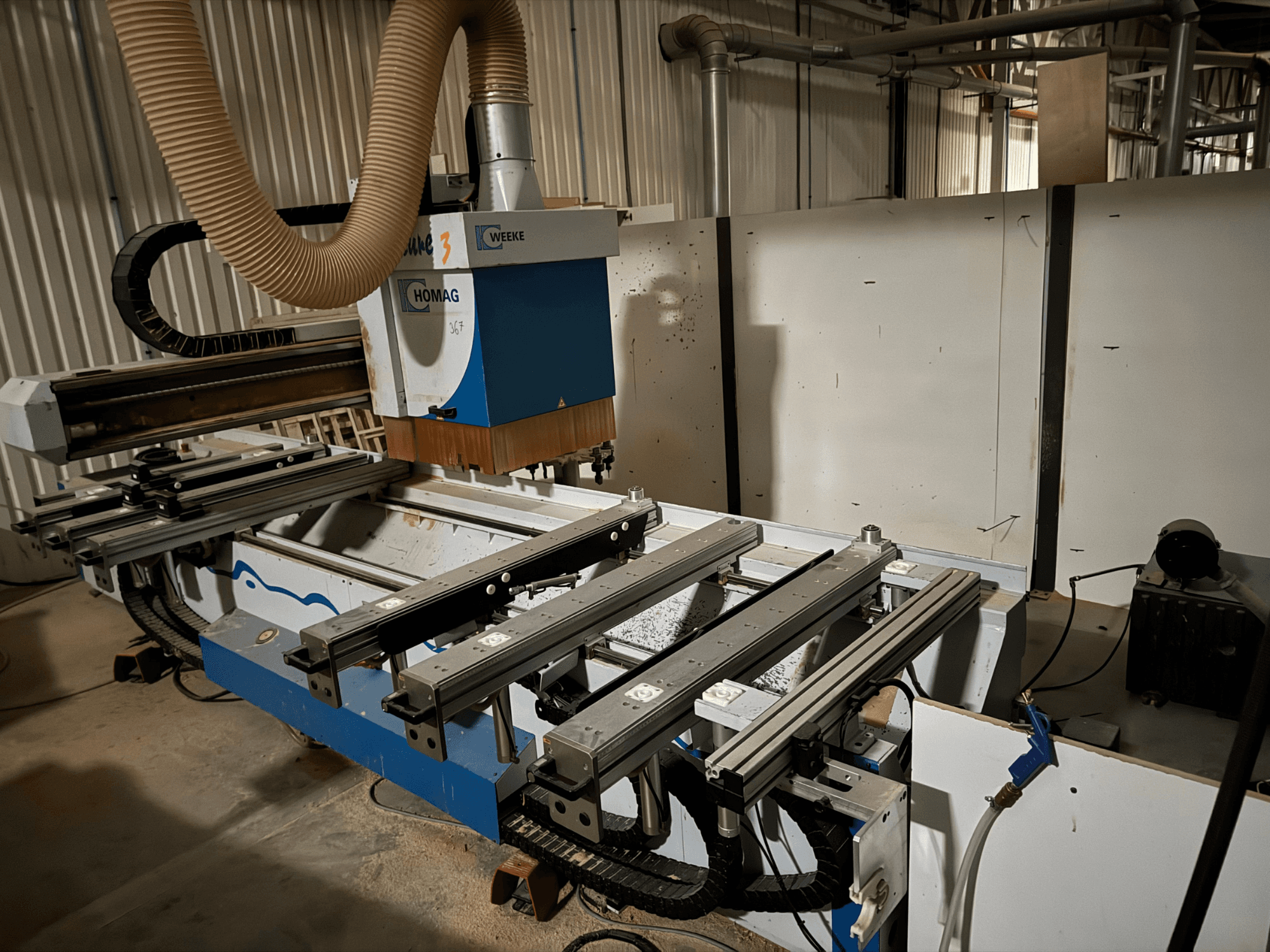 Maszyna CNC HOMAG o niebiesko-białej konstrukcji, widok z boku, wyposażona w przesuwne szyny, system odsysania pyłu i zamontowane narzędzia.
