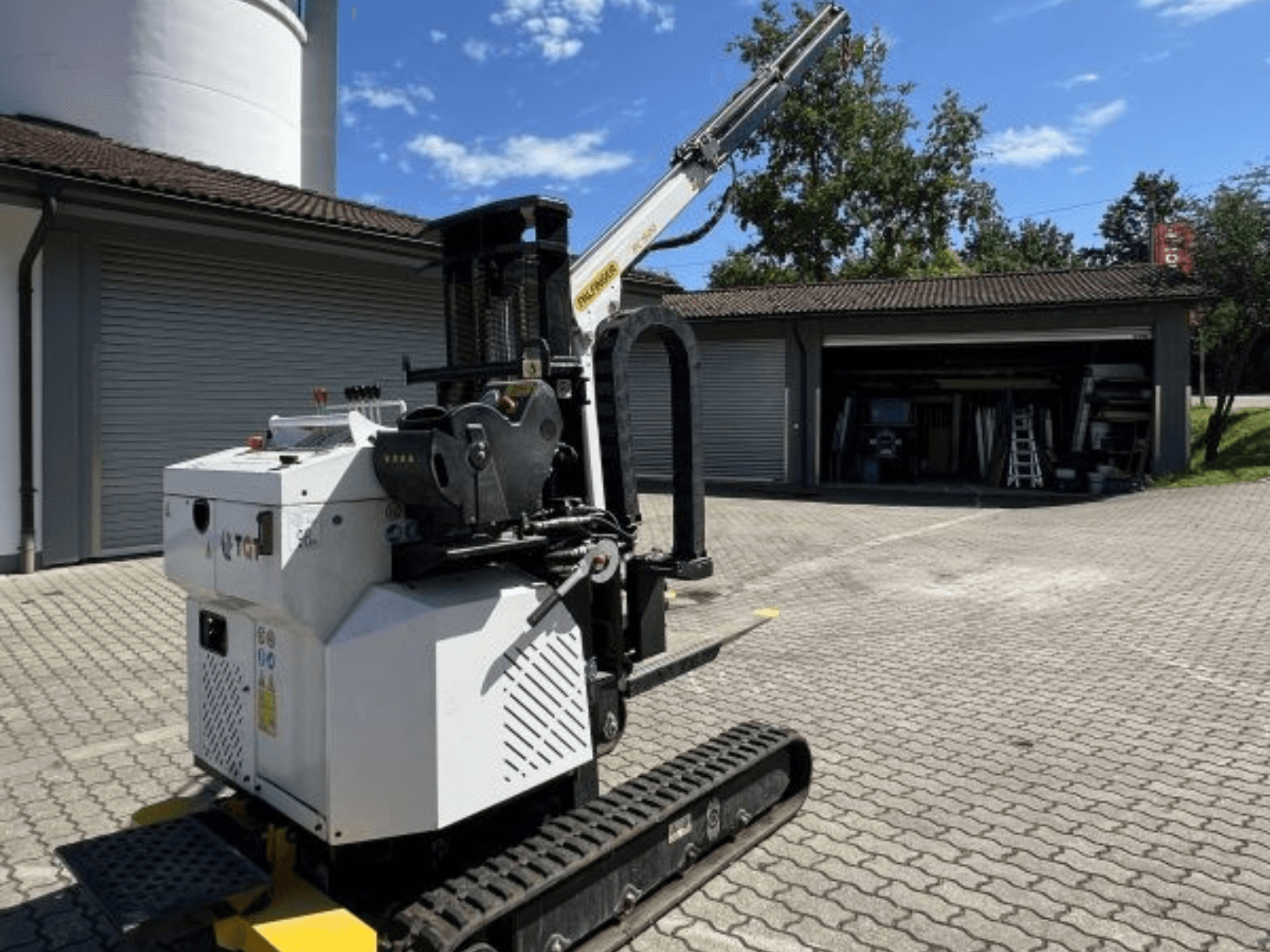 Mini żuraw budowlany TGI, widok z boku, pokazujący gąsienice i zamontowany wysięgnik, zaparkowany na ceglanej powierzchni przy garażu.