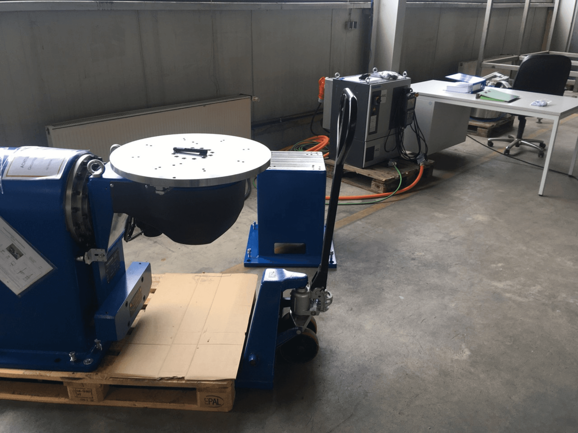 Niebieski sprzęt przemysłowy z okrągłą podstawą i panelem sterowania, widziany z przodu, na drewnianej palecie w otoczeniu biurowym.