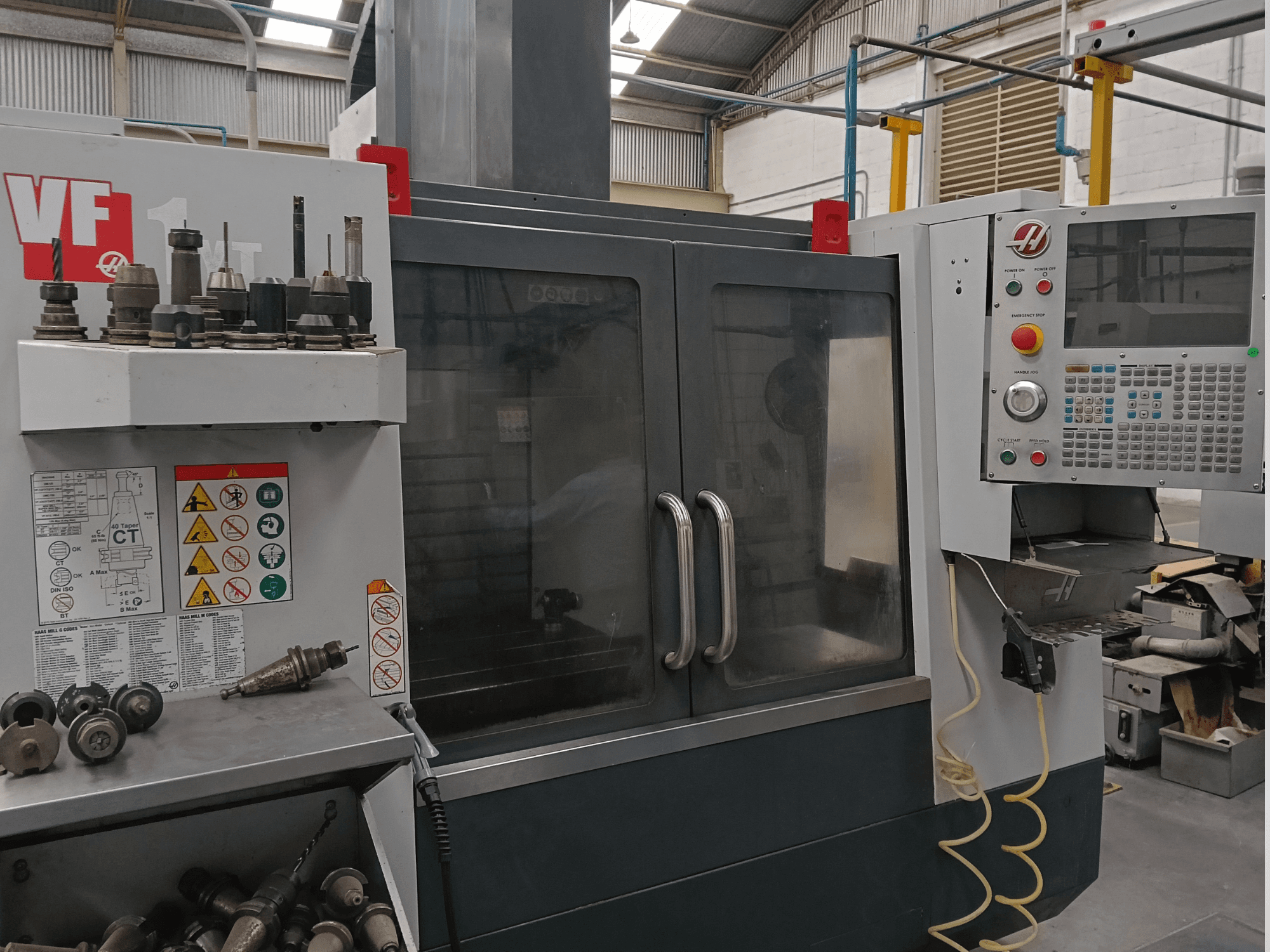 Pionowe centrum obróbcze HAAS VF-1YT-3 widziane z przodu, z widocznymi elementami sterującymi, uchwytami narzędziowymi oraz etykietami bezpieczeństwa.