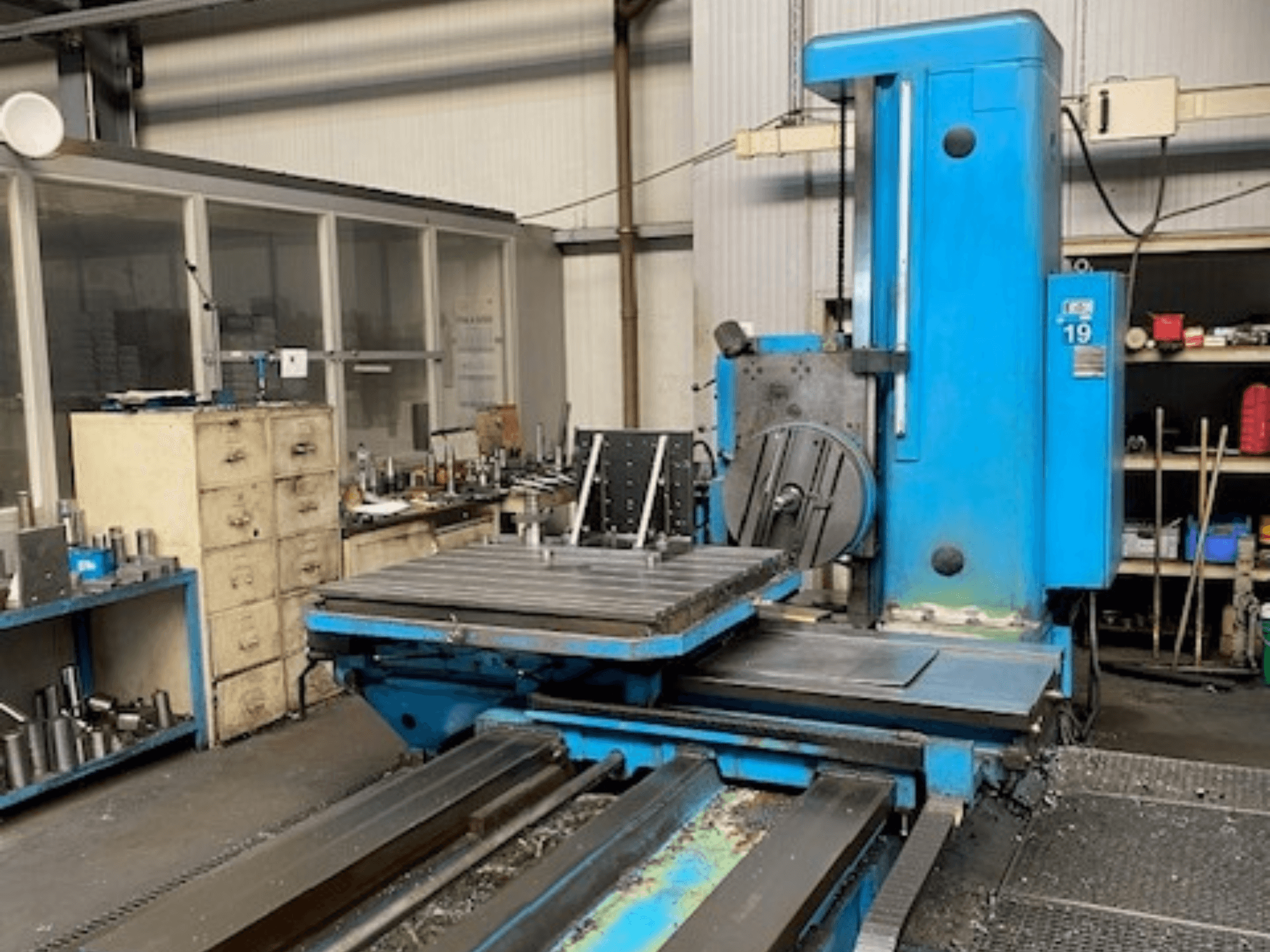 Niebieska frezarka w widoku z przodu, model 19, na podłodze warsztatu z narzędziami i szafką w tle.