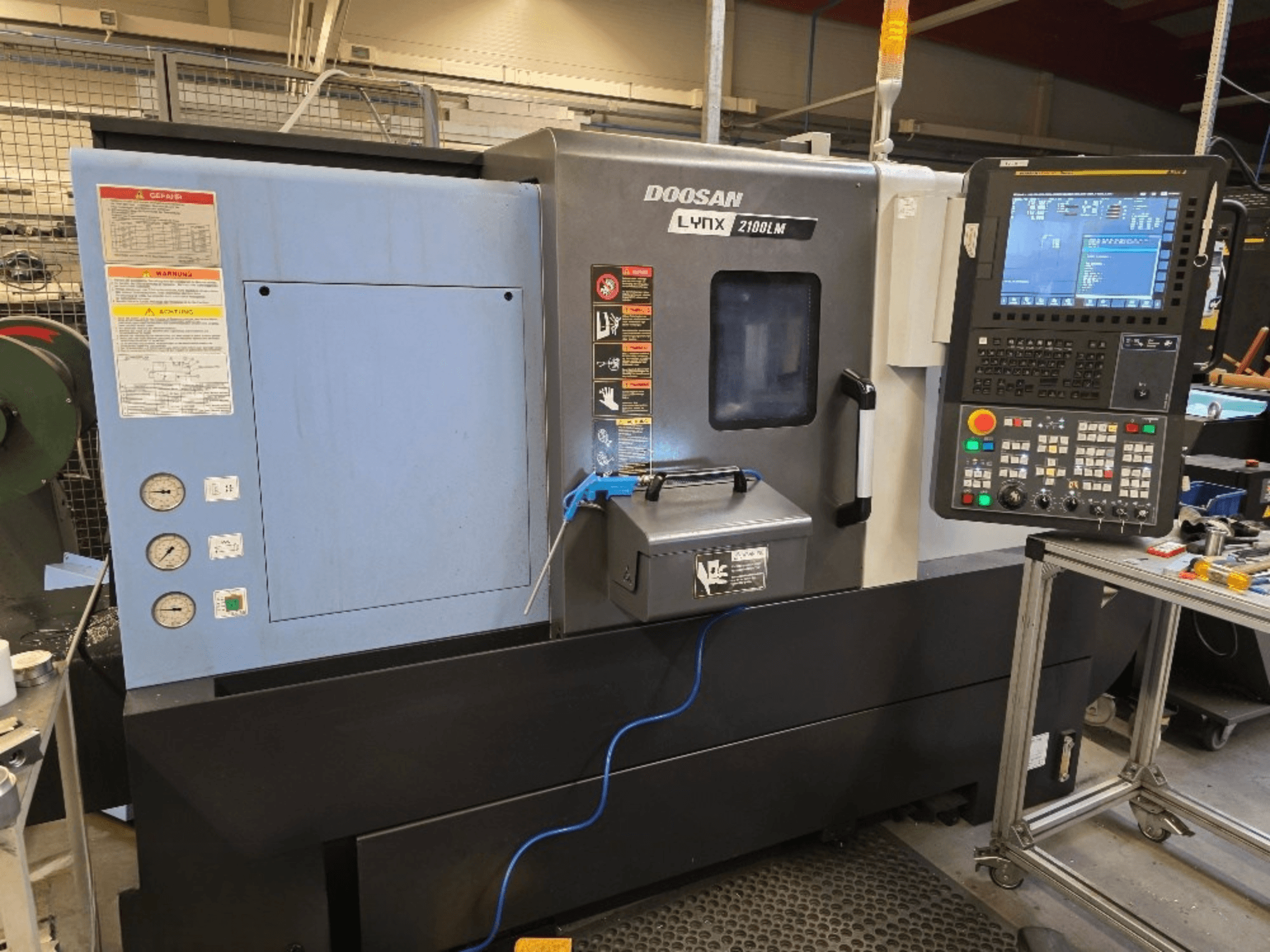 Tokarka CNC Doosan Lynx 2100LM, widok z przodu, z panelem sterowania, manometrami i ostrzeżeniami dotyczącymi bezpieczeństwa.