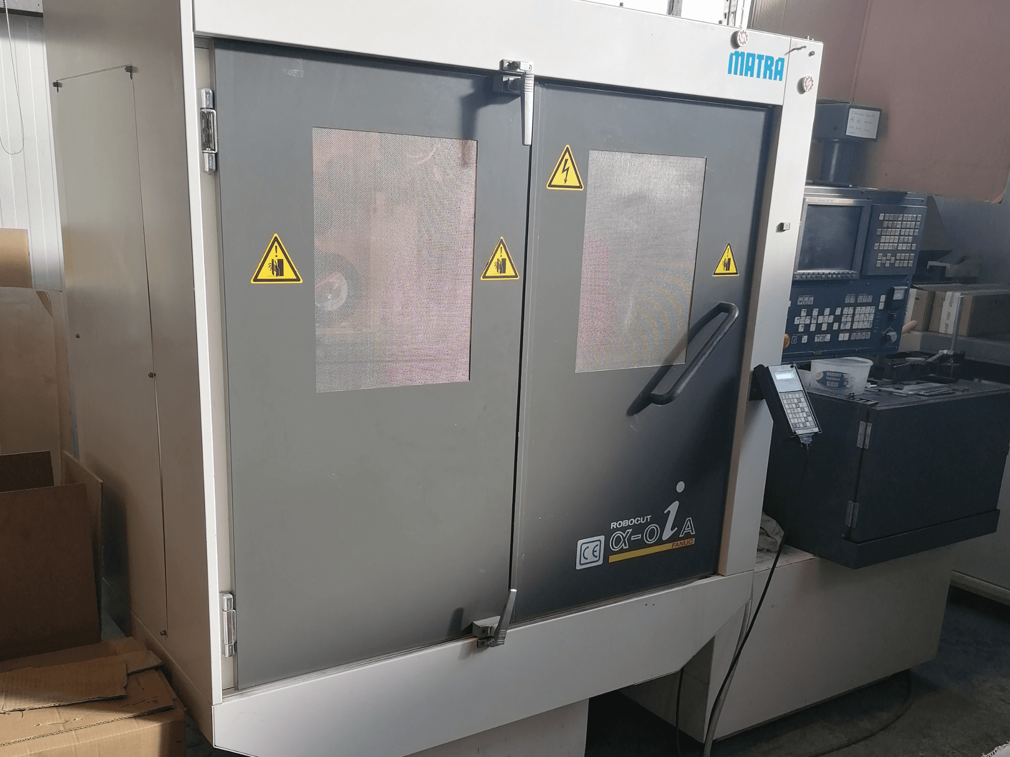 Maszyna Robocut α-oZα firmy Matra, widok z przodu pokazujący ostrzeżenia bezpieczeństwa, panel sterowania i podwójne drzwi z siatkowymi oknami.