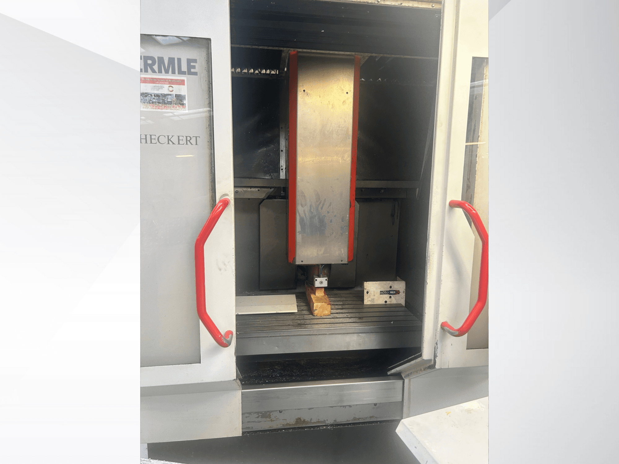 Widok z przodu maszyny CNC Hermle z metalicznym wnętrzem, czerwonymi uchwytami i obrabianym przedmiotem na stole.