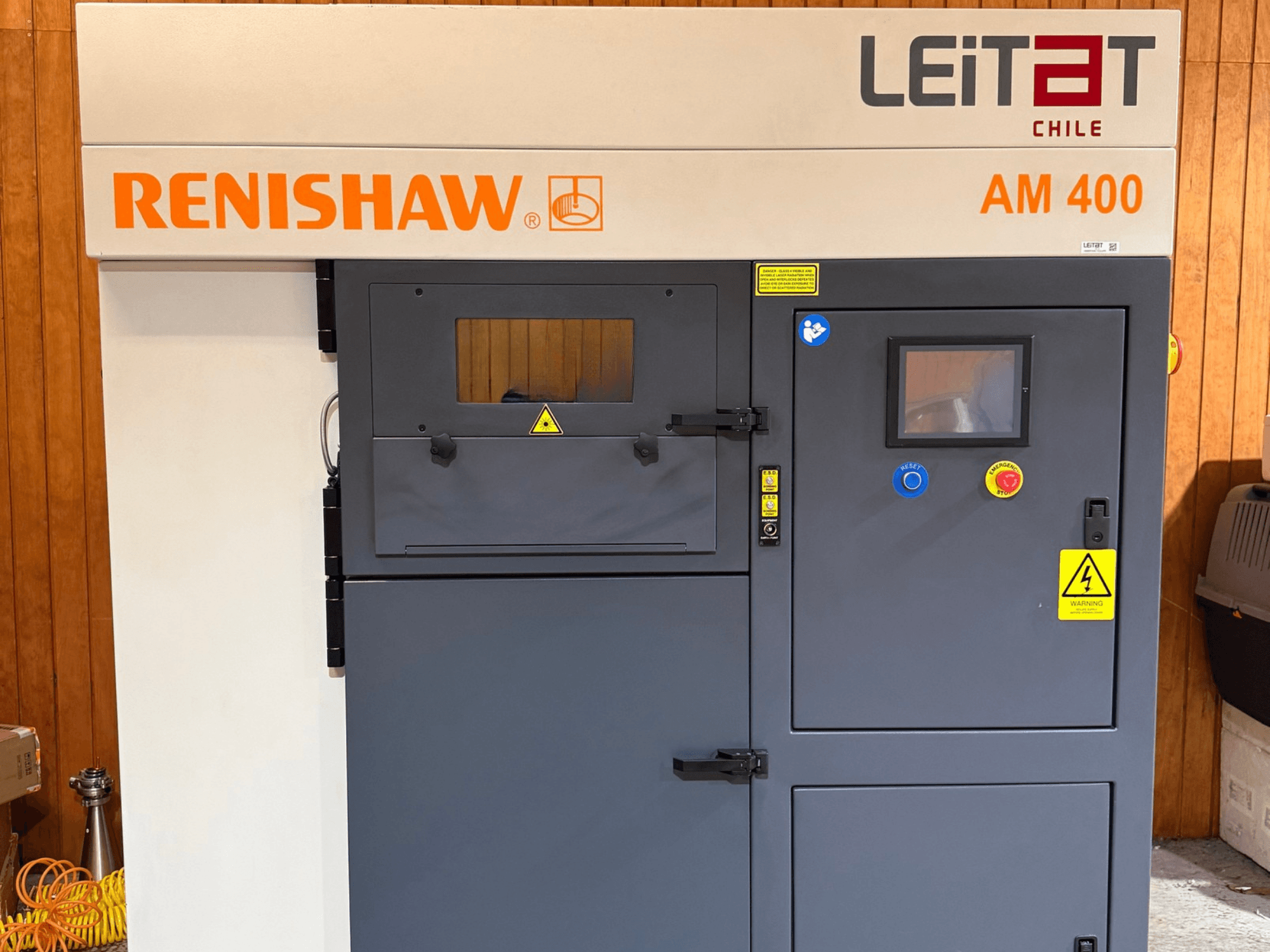 Widok z przodu metalowej drukarki 3D RENISHAW AM400, przedstawiający panel sterowania, etykiety ostrzegawcze oraz otwory serwisowe.
