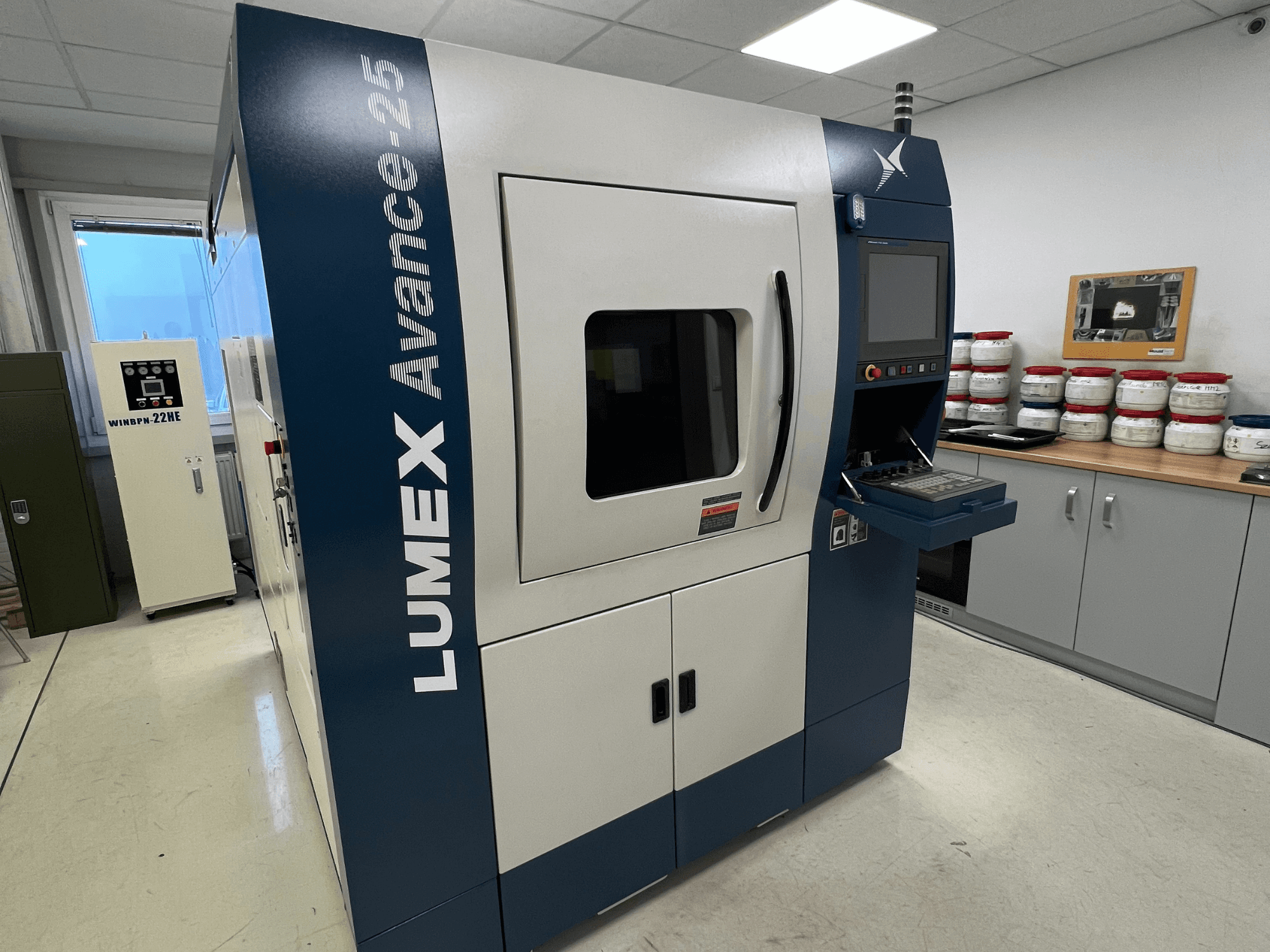 Drukarka 3D LUMEX Avance-25 w warunkach laboratoryjnych, widok z przodu. Panel sterowania z widocznym ekranem i przyciskami, sprzęt w tle.