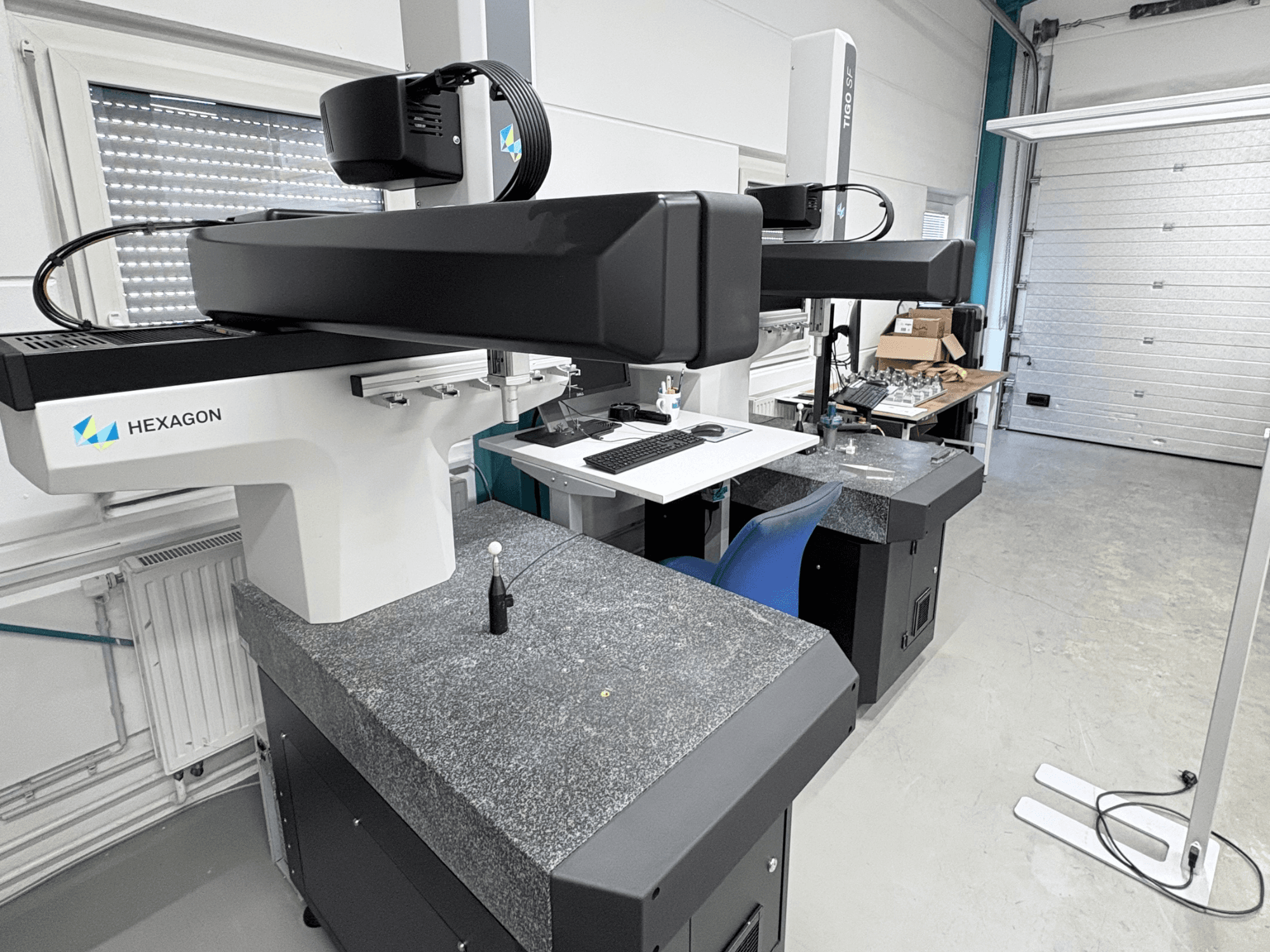 Maszyna CMM Hexagon Tigo SF w warsztacie, wyposażona w granitową podstawę, sondę pomiarową oraz znajdującą się w pobliżu stację roboczą z narzędziami.