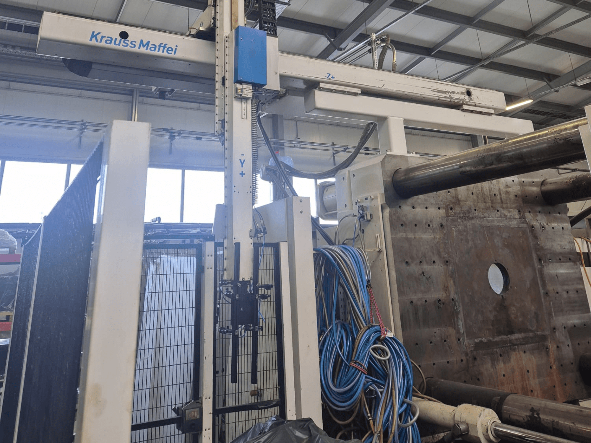 Hydrauliczna wtryskarka Krauss Maffei 1300-12000 MX, widok z przodu z ramionami mechanicznymi i przyłączami hydraulicznymi.