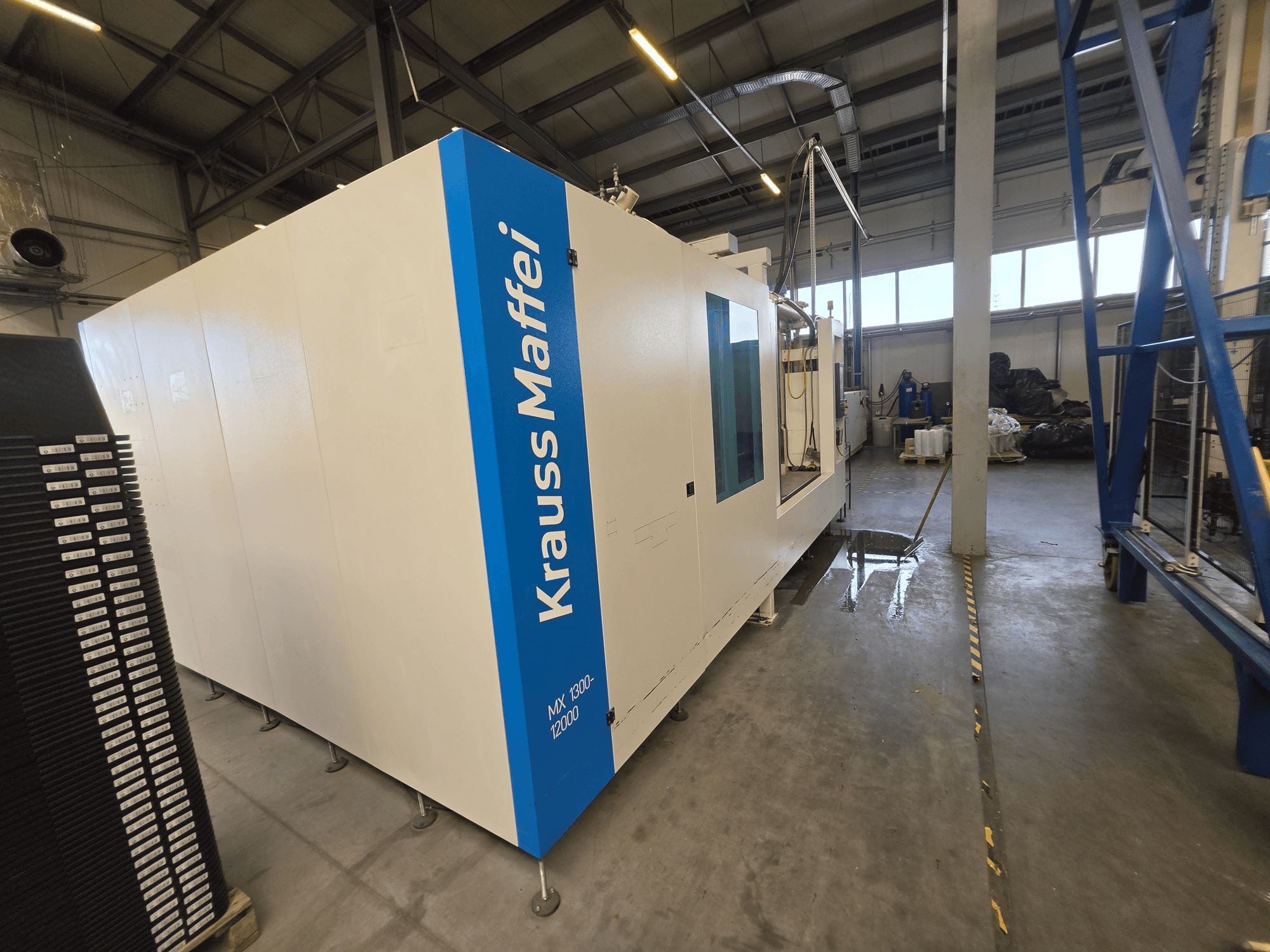 Hydrauliczna wtryskarka Krauss Maffei MX 1300-12000 widziana z boku, z widocznym dużym białym podwoziem i niebieskimi oznaczeniami firmowymi.