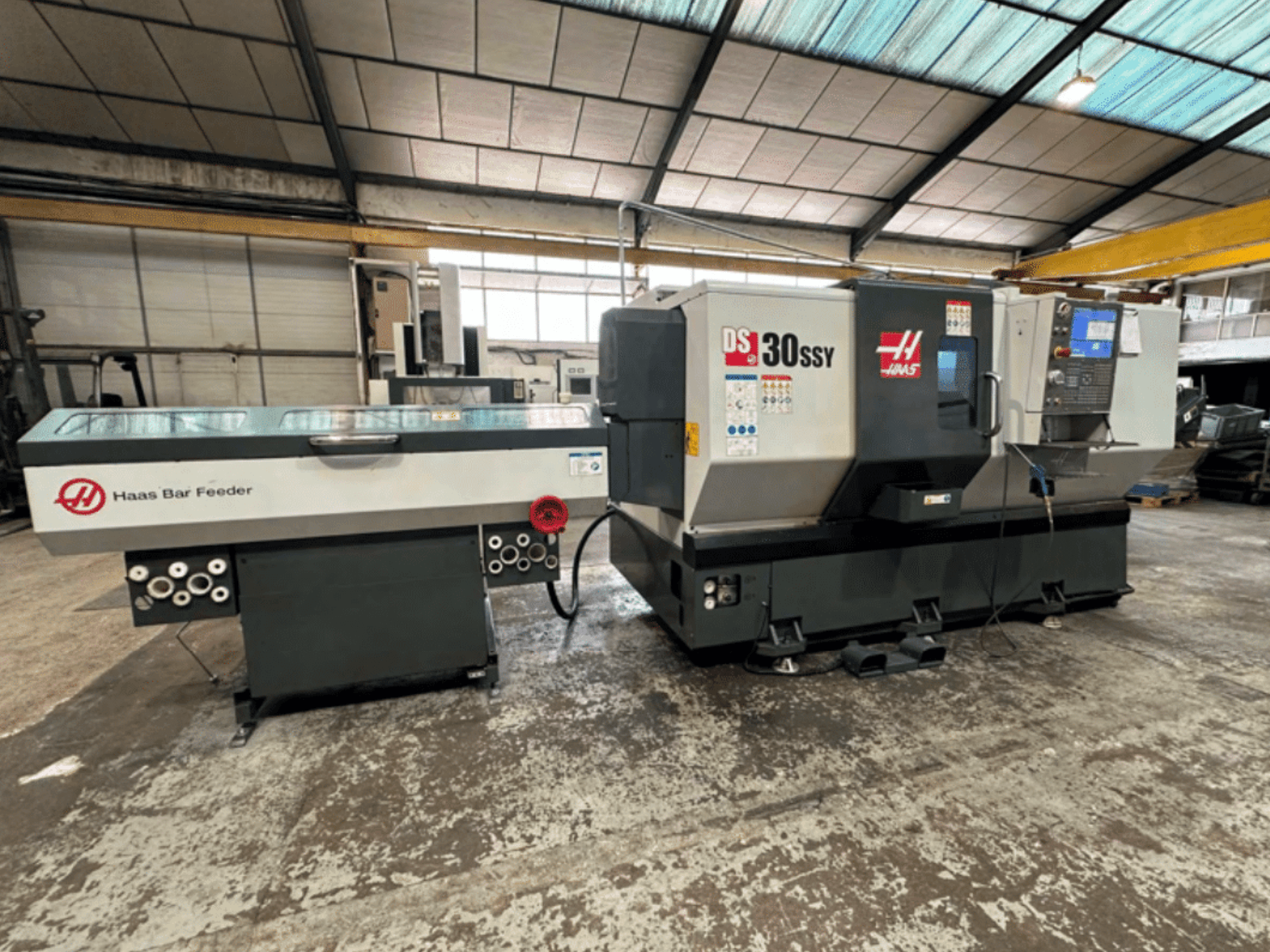 Tokarka CNC Haas DS-30SSY z podajnikiem prętów Haas, widok z boku przedstawiający panel sterowania i mechanizm podawania w warunkach warsztatowych.