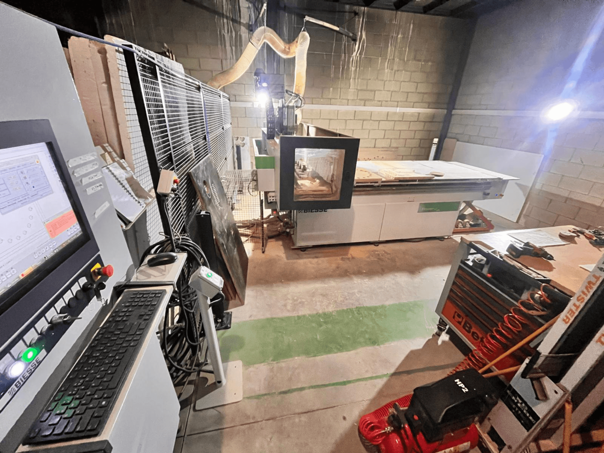 Maszyna CNC Biesse w warsztacie, widok z boku pokazujący panel sterowania, odsysanie pyłu, narzędzia i różne materiały na stole.