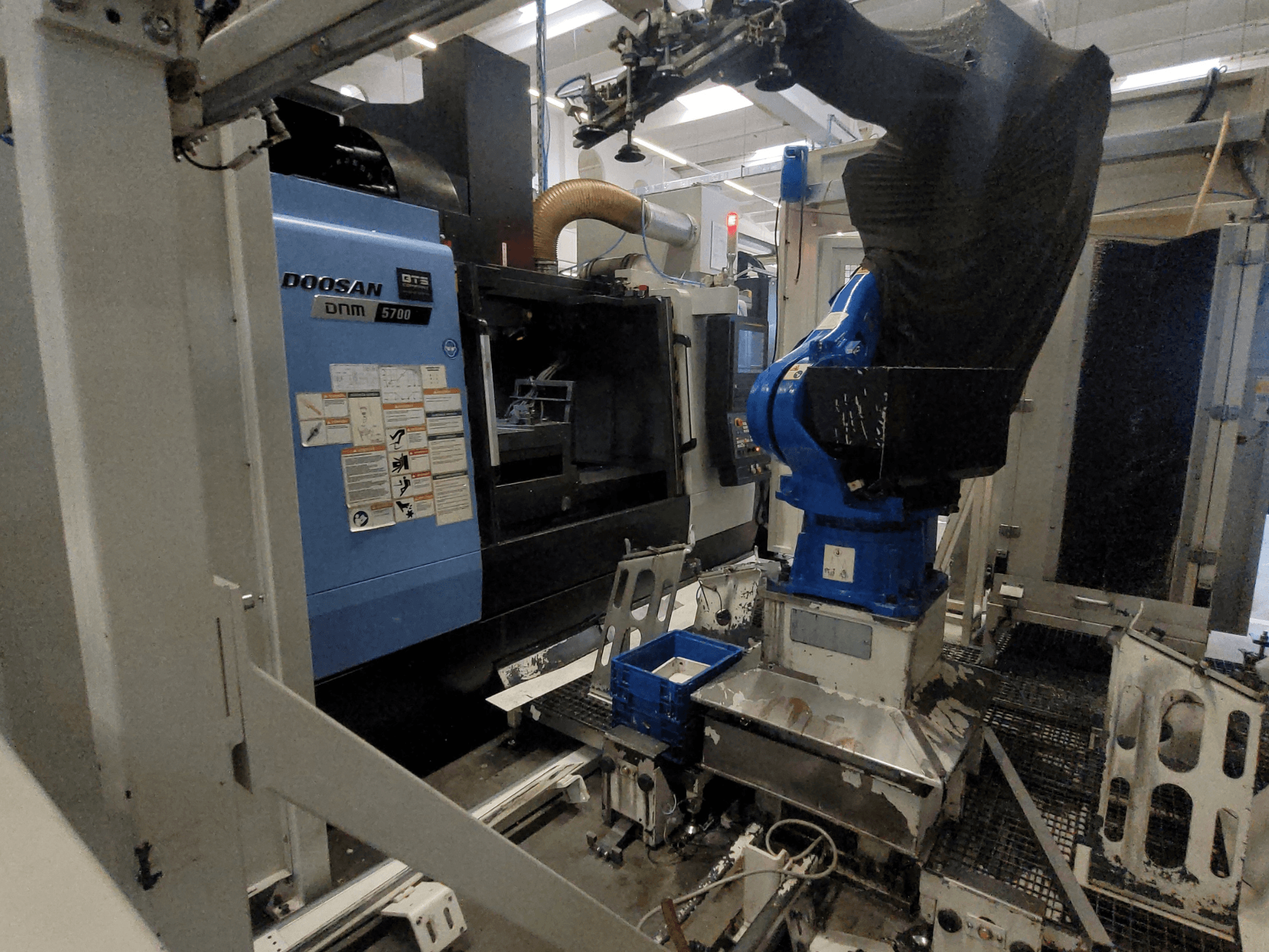 Pionowe centrum obróbcze DOOSAN DNM 5700 w warunkach fabrycznych, przedstawiające widok z przodu oraz zintegrowane ramię robota.