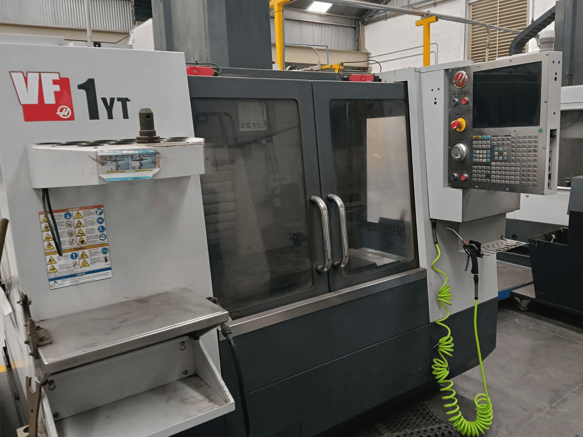 Pionowe centrum obróbcze HAAS VF-1YT-2, widok z przodu, przedstawiający panel sterowania, uchwyt narzędziowy, etykiety bezpieczeństwa oraz obszar obróbki.