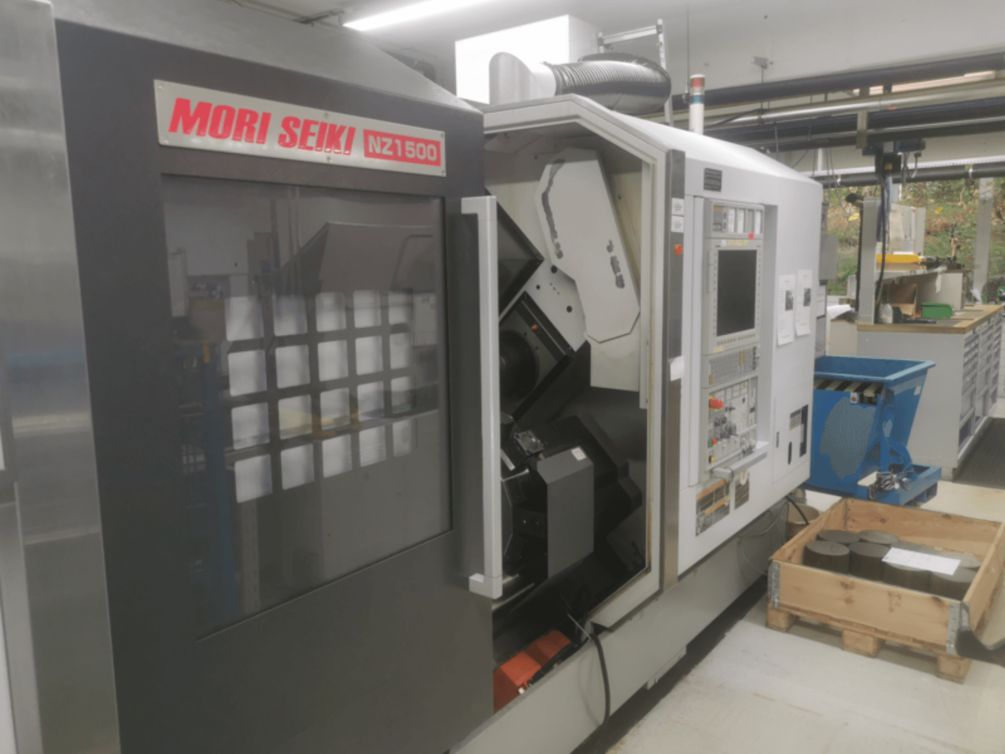 Maszyna CNC MORI SEIKI NZ1500 pod niewielkim kątem, pokazująca panel sterowania i obszar roboczy z metalowymi elementami na drewnianej palecie.