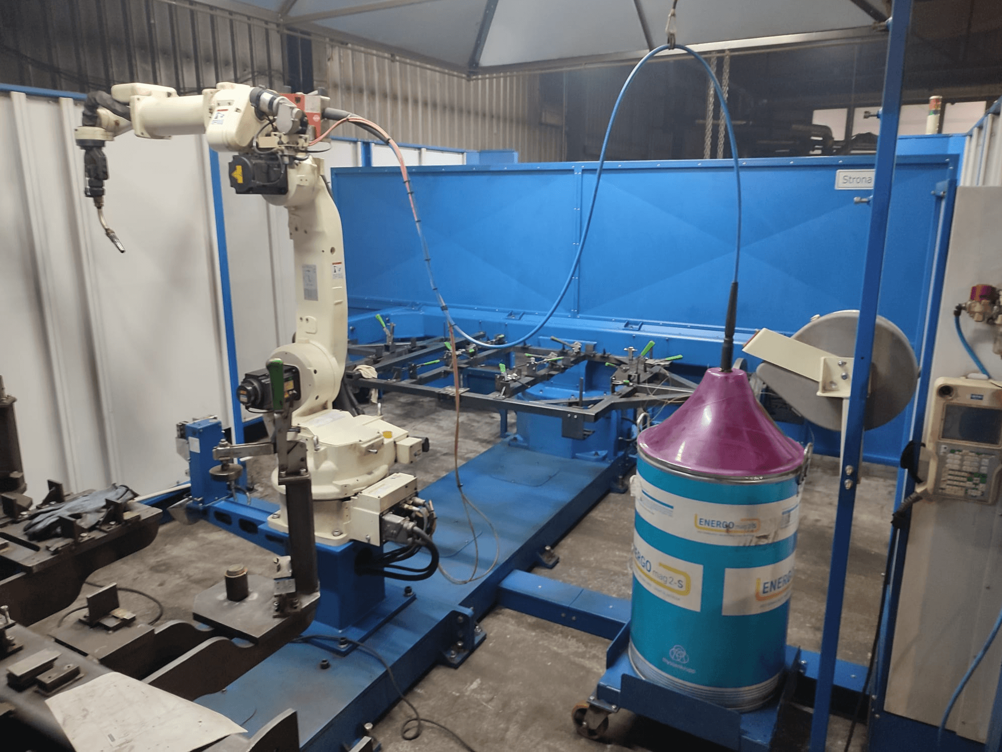 Przemysłowe ramię robota z biało-niebieską podstawą, skonfigurowane w warsztacie z niebieską powierzchnią roboczą i metalowymi elementami.