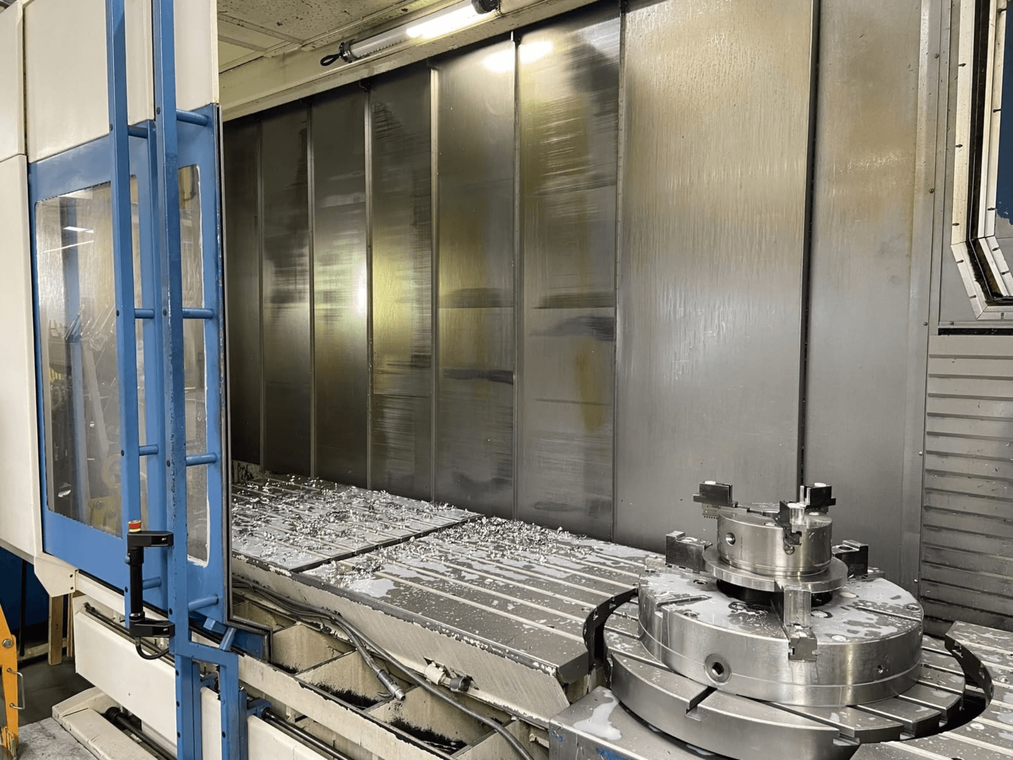 Pionowe centrum obróbcze AXA VHC-3-3000-M/E, widok z przodu, przedstawiający wióry metalowe na stole roboczym oraz narzędzia skrawające.