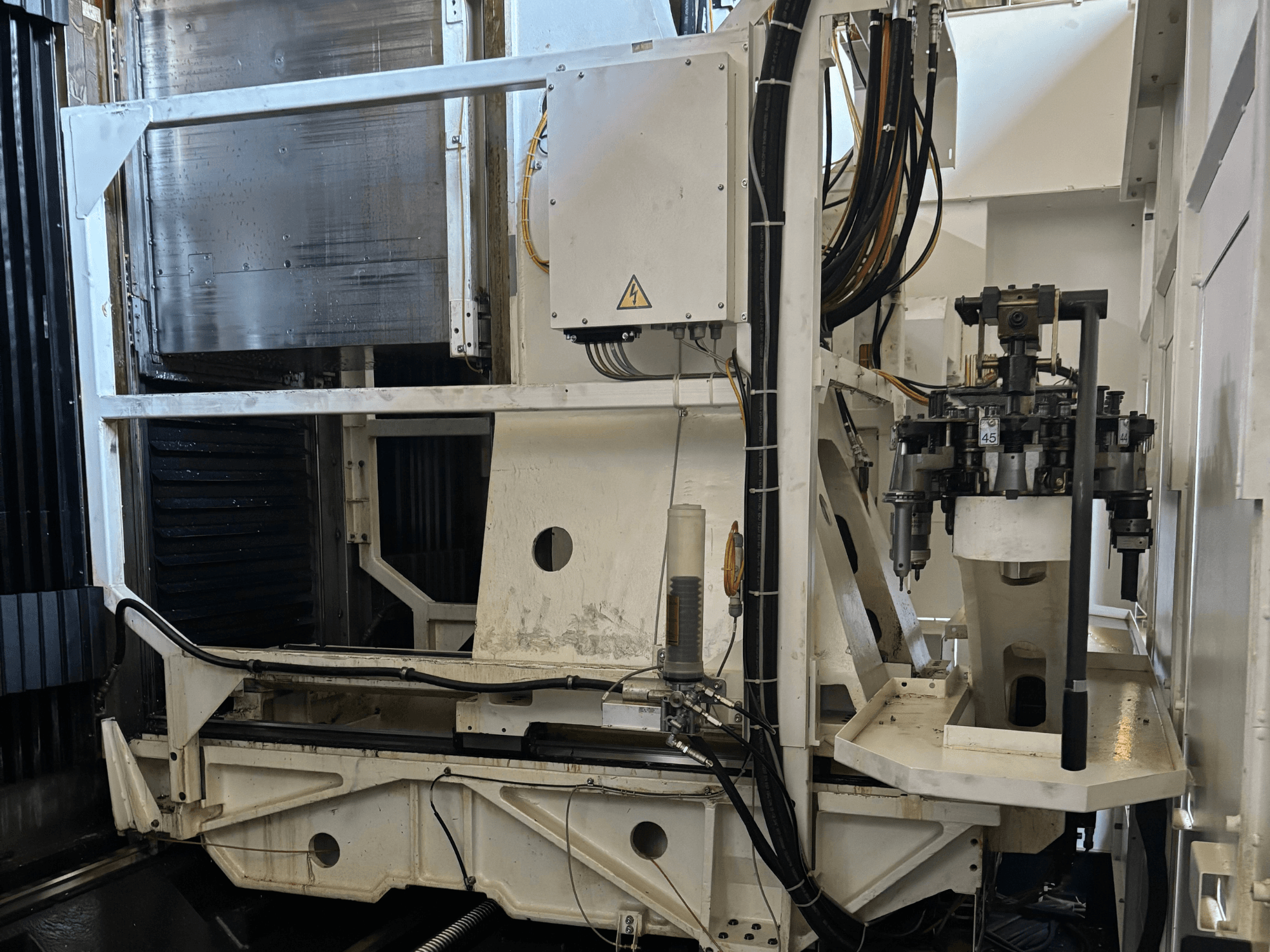 Widok z boku maszyny przemysłowej przedstawiający białą frezarkę CNC z widocznymi narzędziami, rurami i panelem sterowania.