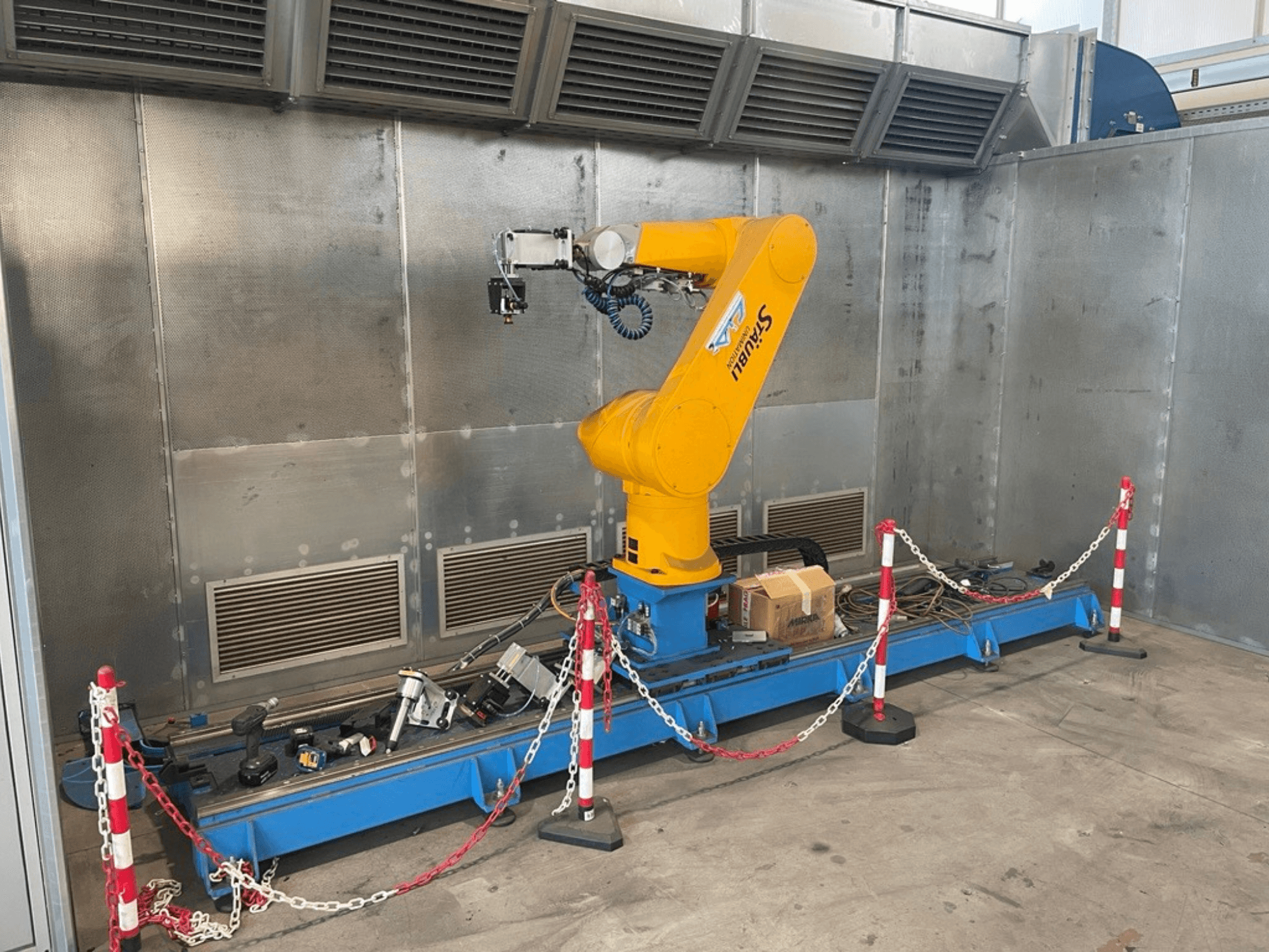 Ramię robota STAUBLI CS8C-RX160 w warsztacie, widok z boku, z kablami i różnymi narzędziami na niebieskiej podstawie otoczonej barierami ochronnymi.