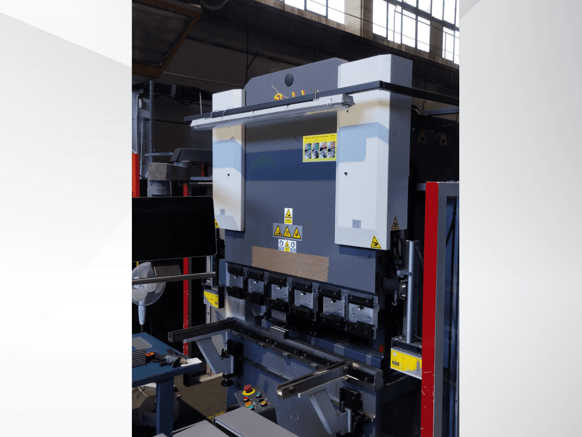 Prasa krawędziowa, widok z przodu. Posiada widoczne elementy sterujące, naklejki bezpieczeństwa i wiele uchwytów matryc do precyzyjnego gięcia.