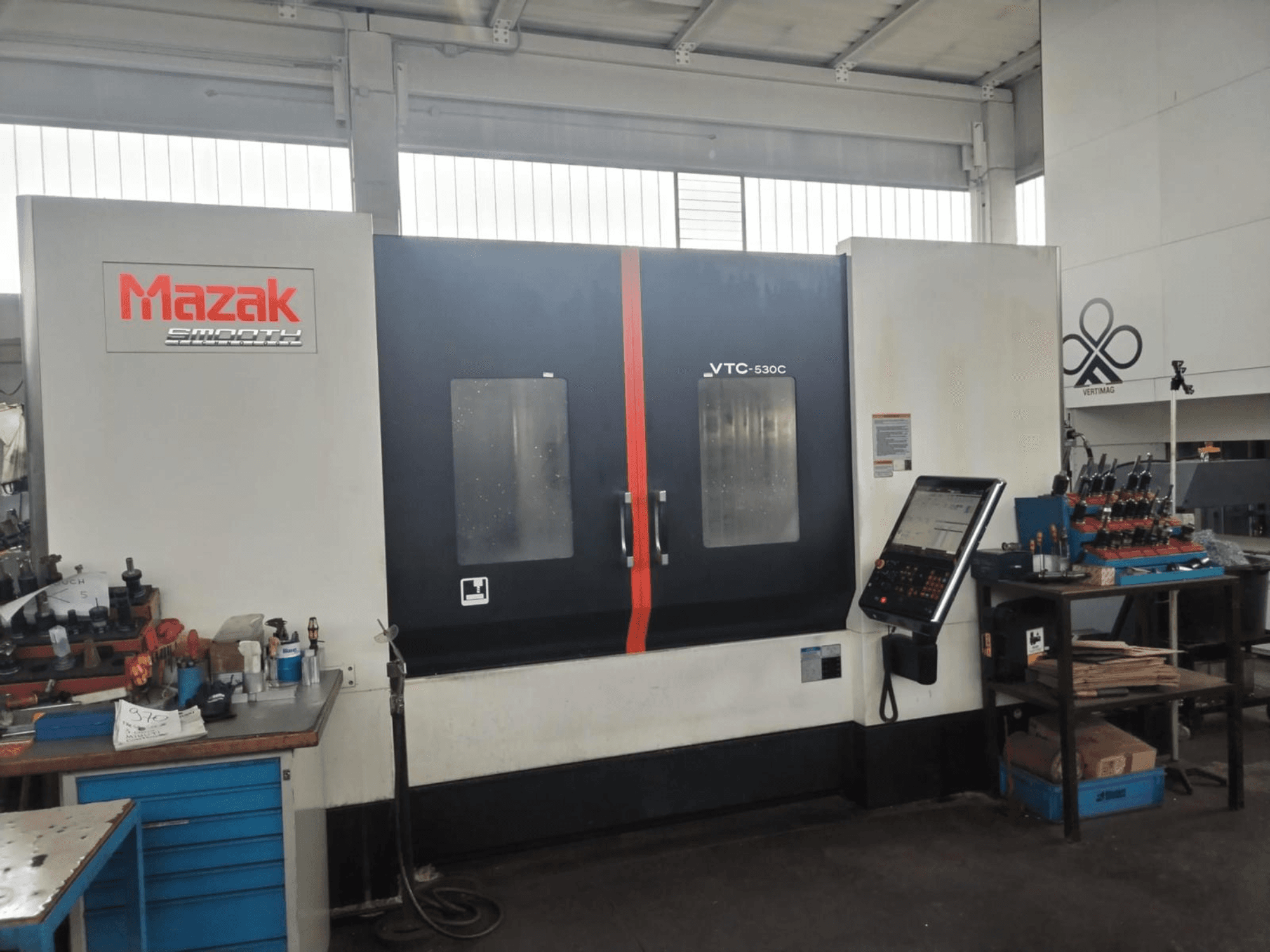 Pionowe centrum obróbcze Mazak VTC-530C w warsztacie, wyposażone w panel sterowania i narzędzia umieszczone na bocznym stole.