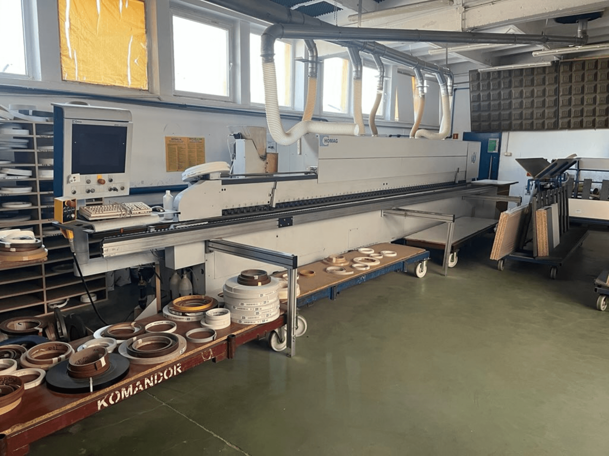 Okleiniarka HOMAG w warsztacie, z elementami sterującymi i różnymi materiałami do oklejania krawędzi na niebieskim wózku. Widok z boku.