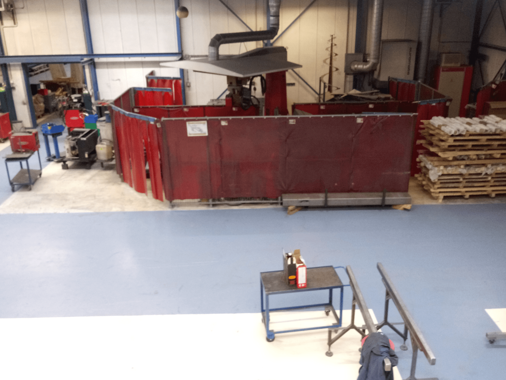 Przemysłowa przestrzeń robocza z czerwonymi kurtynami spawalniczymi, maszynami i paletami; widok z góry przedstawiający układ i wyposażenie.