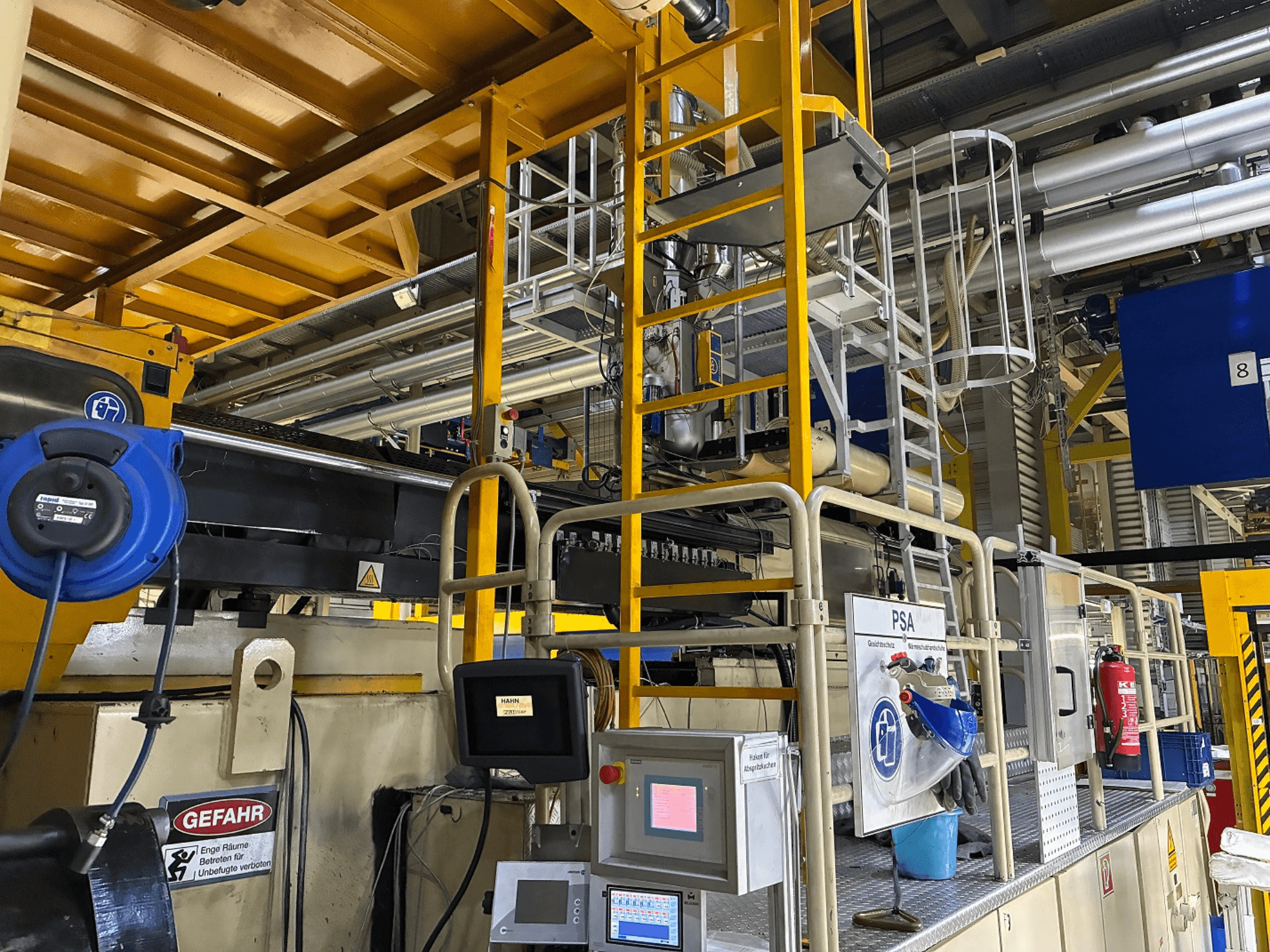 Panel sterowania maszyny przemysłowej z niebieskim sprzętem ochronnym i żółtym rusztowaniem, widok z boku pokazujący rury i znaki ostrzegawcze.