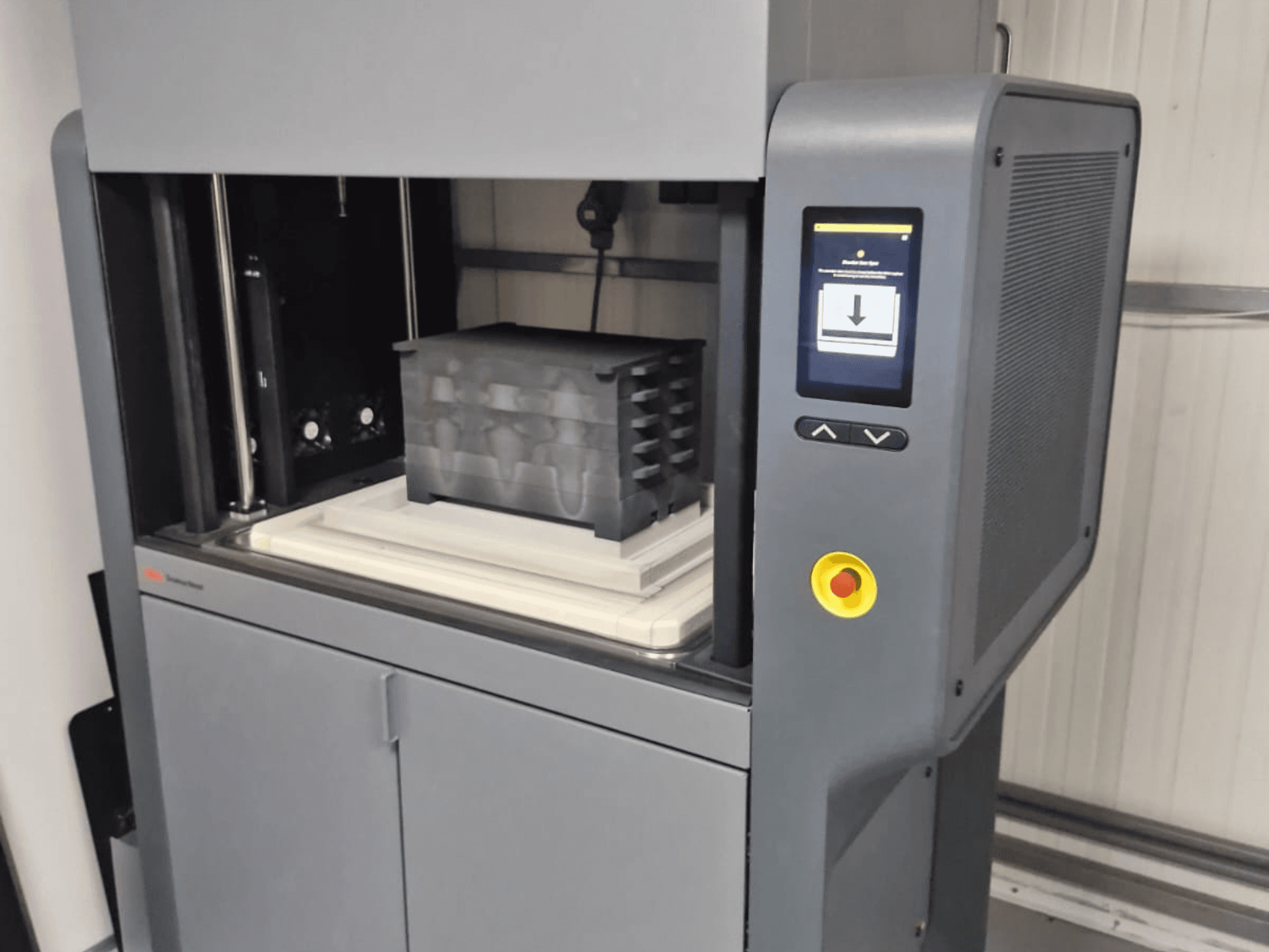 Drukarka 3D Metal Studio 2 firmy Desktop Metal, widok z przodu przedstawiający panel sterowania i stół roboczy z czarnym pojemnikiem.