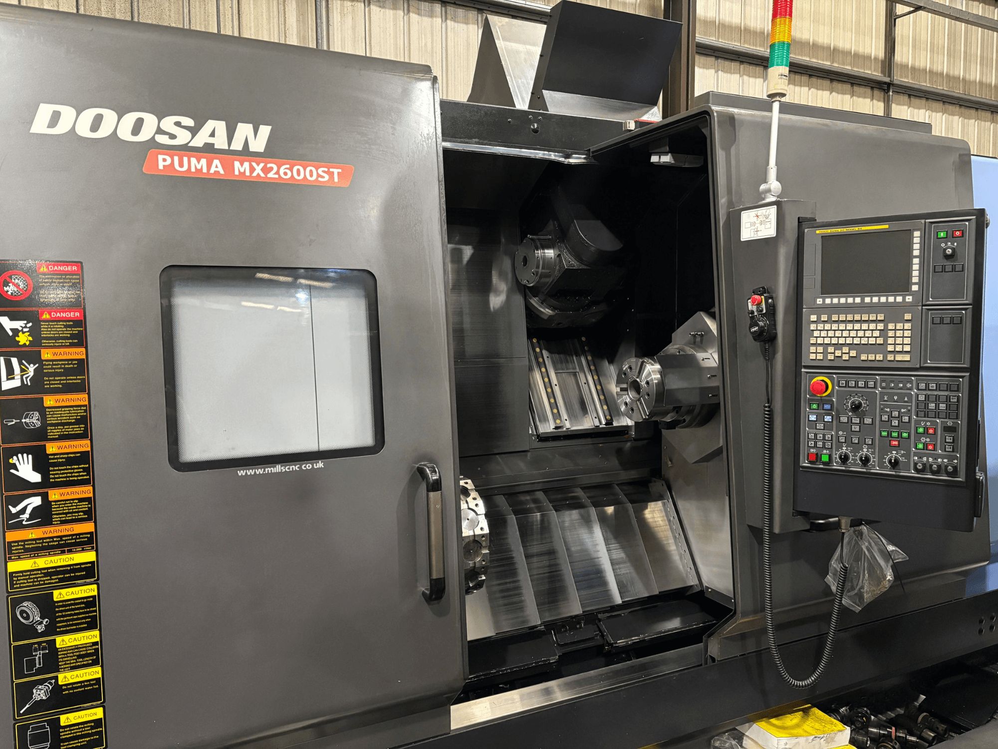 Tokarka CNC Doosan PUMA MX2600ST, widok z przodu, pokazujący panel sterowania, wrzeciono i obszar obróbki z widocznymi ostrzeżeniami dotyczącymi bezpieczeństwa.