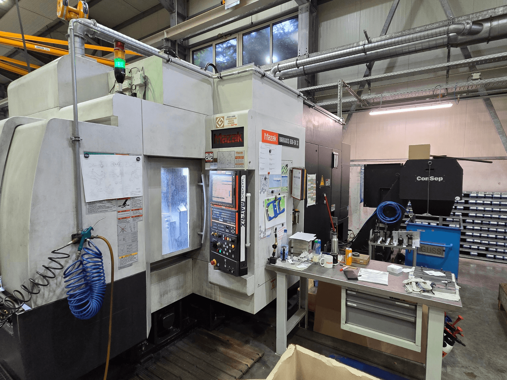 Maszyna CNC Mazak Variaxis 630-5X w warsztacie, widok z przodu z panelem sterowania, wężem i akcesoriami na bocznym stole.