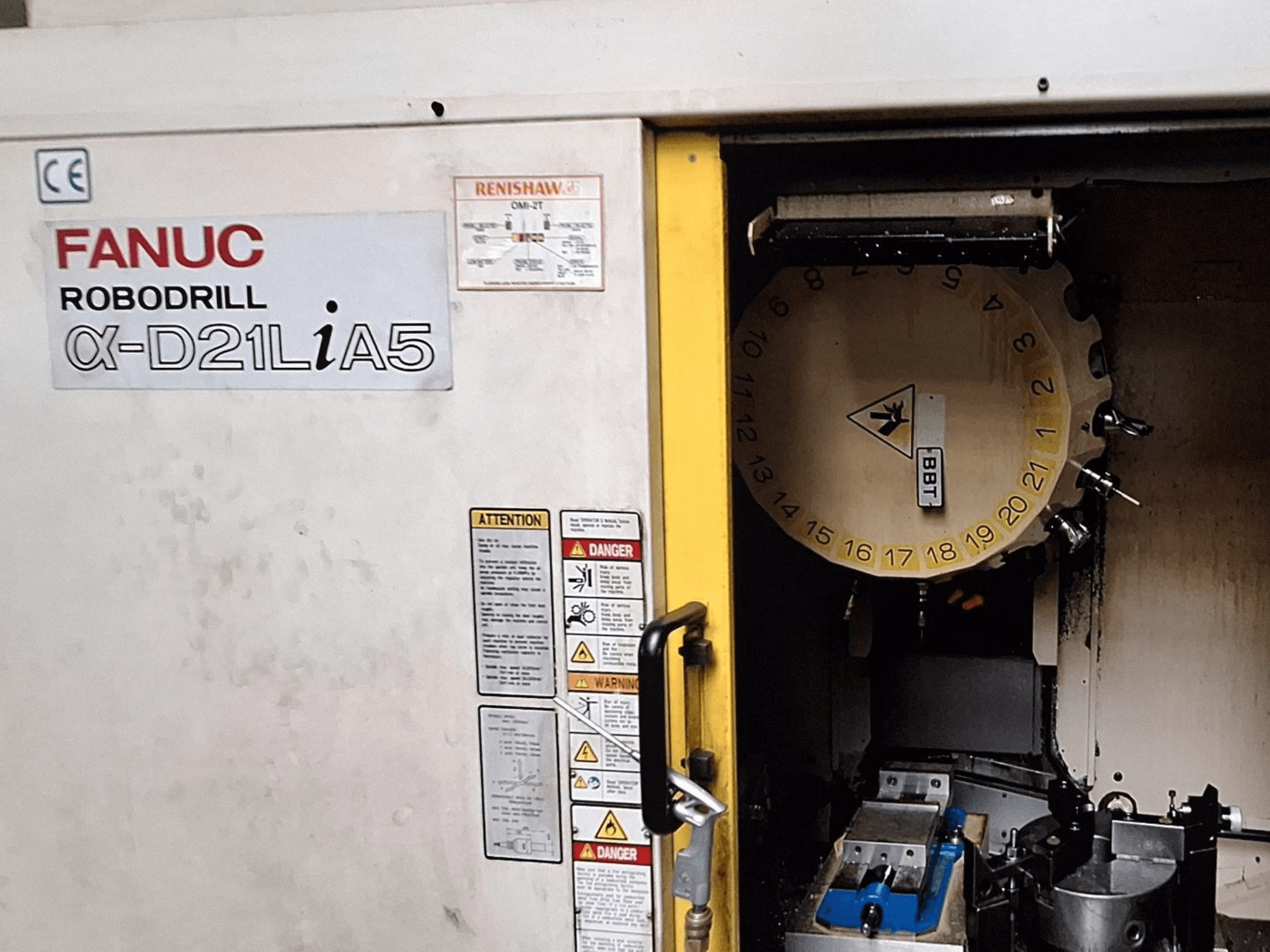 FANUC Robodrill α-D21LiA5, widok z przodu. Pokazuje karuzelę narzędziową i obszar obróbki z zaciskami i ostrzeżeniami na drzwiach.