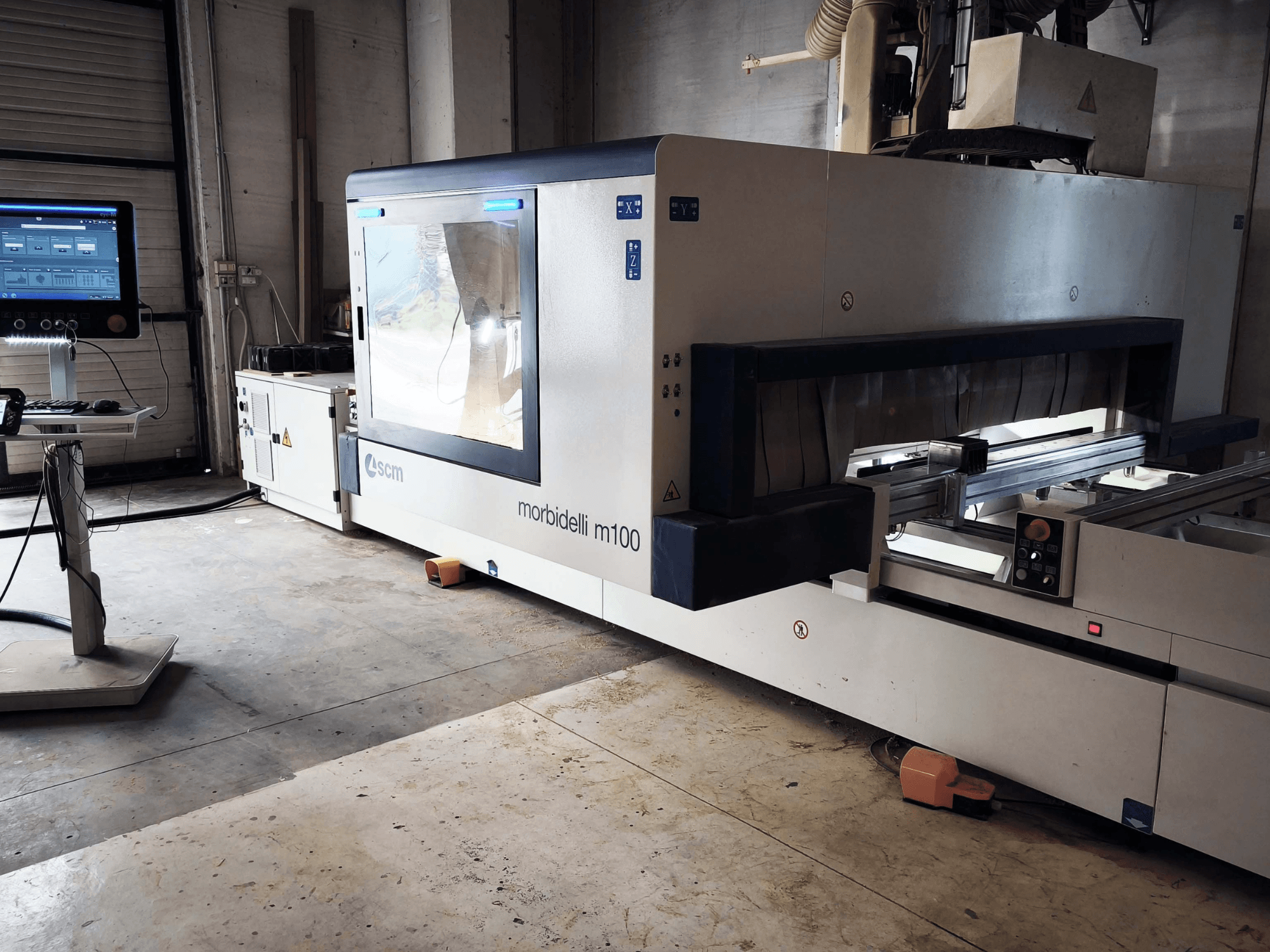 Centrum obróbki drewna SCM Morbidelli M 100 CNC widziane z boku, z widocznym panelem sterowania, obszarem roboczym i elementami zabezpieczającymi.