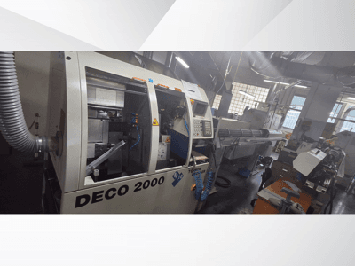 Maszyna CNC DECO 2000 w warsztacie, widok z przodu, elementy sterujące i konfiguracja długiego przedmiotu obrabianego po prawej stronie.