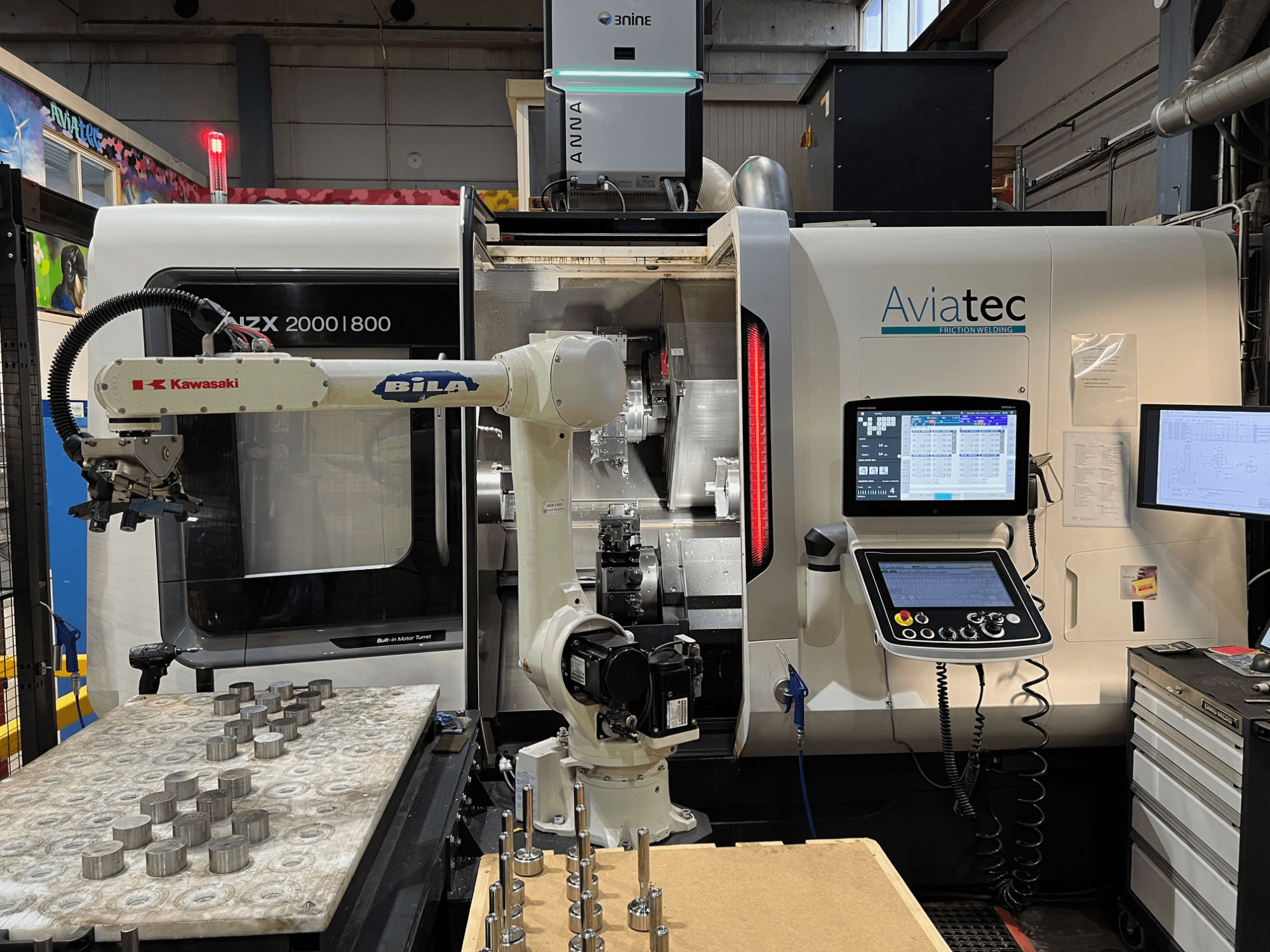 Pozioma tokarka DMG MORI NZX 2000/800SY z ramieniem robota firmy Kawasaki, panelem sterowania oraz elementami metalowymi na stole roboczym.