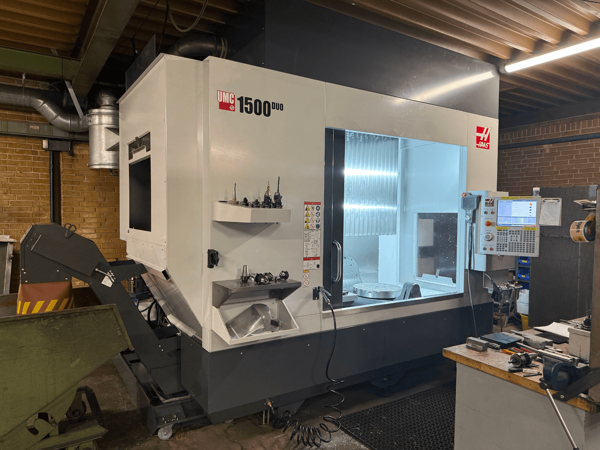 Maszyna CNC Haas UMC 1500DUO, widok z przodu, z dużym oknem, panelem sterowania i uchwytami narzędzi na półce.