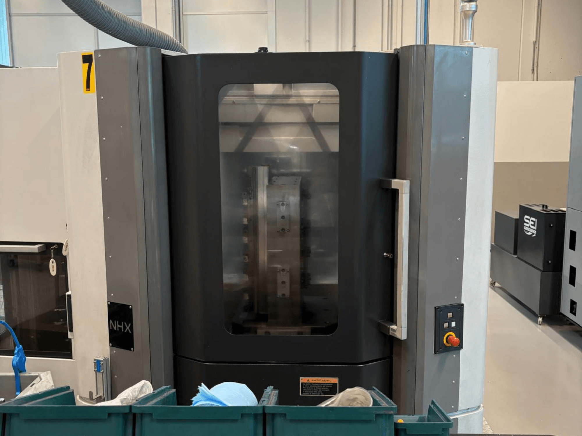 Maszyna CNC NHX w widoku z przodu, pokazująca panel sterowania i czytelny wyświetlacz; umieszczona w warsztacie z zielonymi pojemnikami w pobliżu.