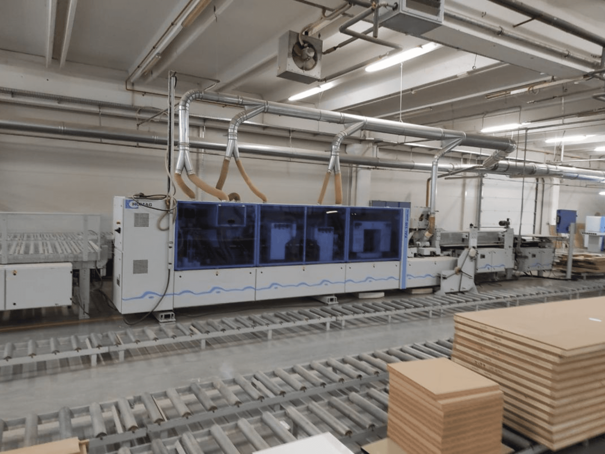 Maszyna produkcyjna HOMAG w fabryce, widok z przodu, z niebieską obudową i systemem przenośników z ułożonymi arkuszami sklejki.